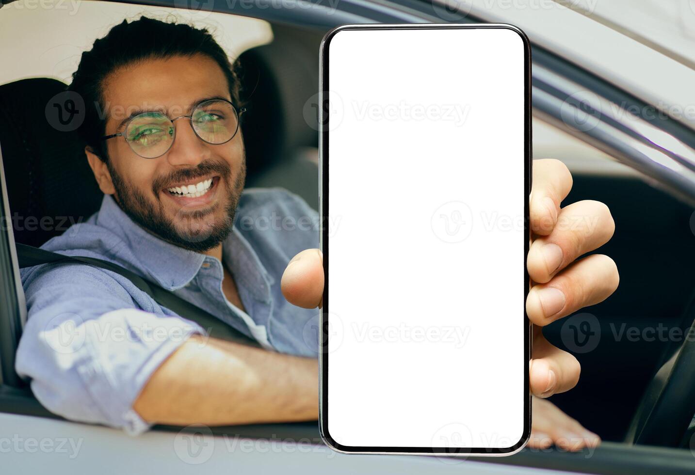 Closeup of young smiling Indian man in glasses showing big mobile phone with white empty screen for mock up sitting on driver seat in new luxury car, holding gadget in hand, selective focus on device photo