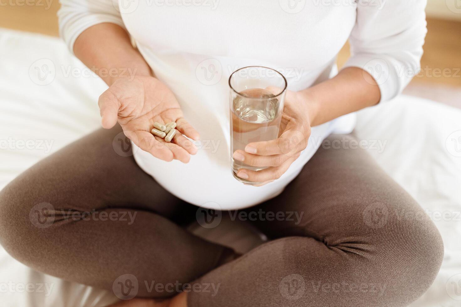 A woman in her third trimester sits comfortably while holding vitamins in one hand and a glass of water in the other. She appears focused on her health and well-being. photo