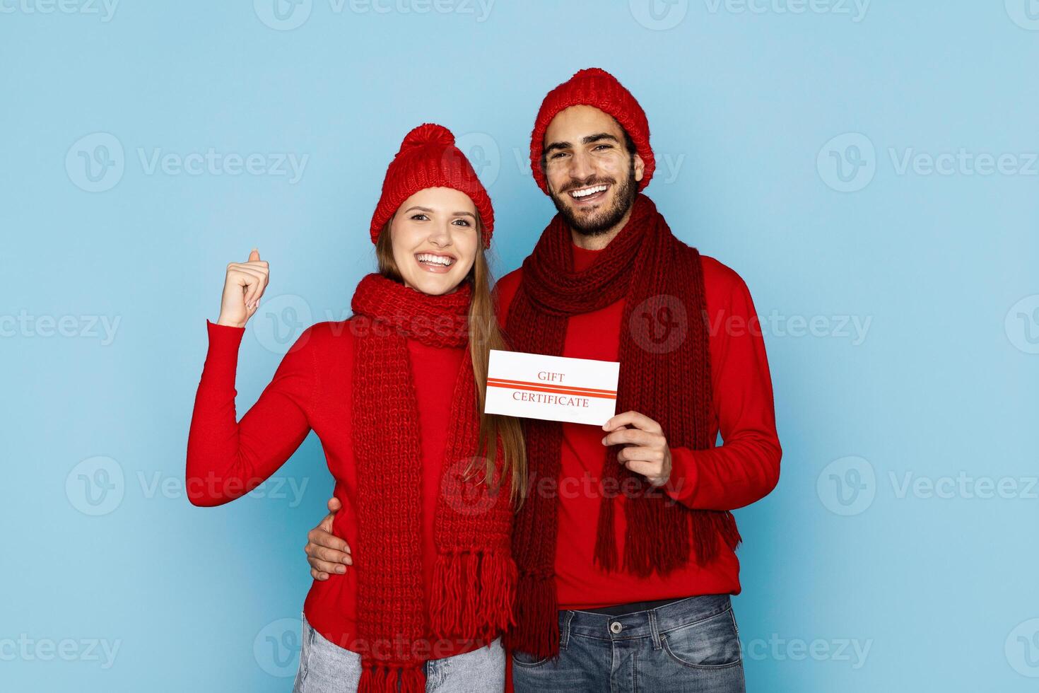 A joyful young couple dressed in matching red hats and scarves holds a gift certificate, smiling against a light blue background, celebrating a special occasion together. photo
