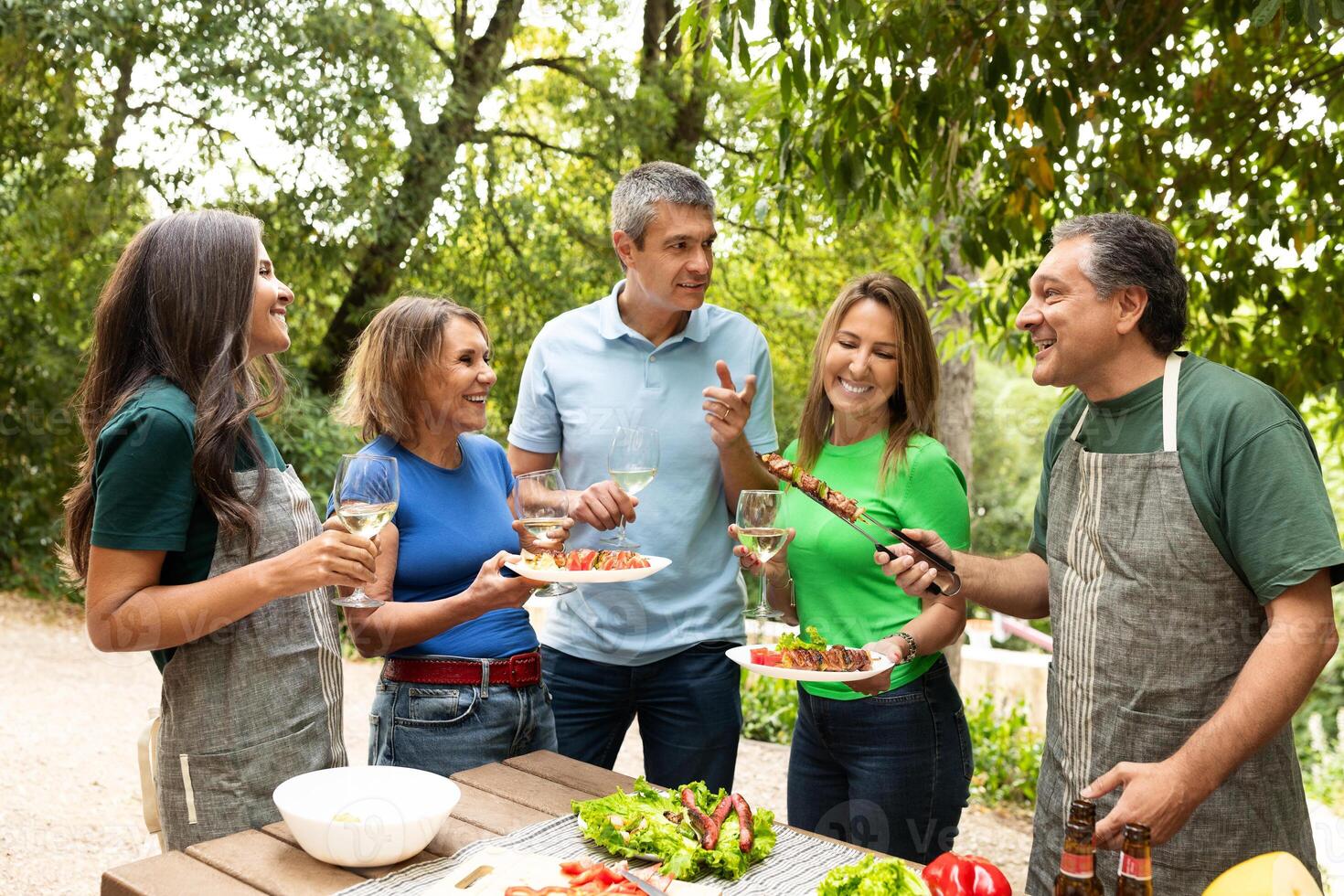 Friends celebrate together at a barbecue, sharing laughter and delicious food in a verdant garden. The sun shines as they toast with drinks and savor the moment. photo