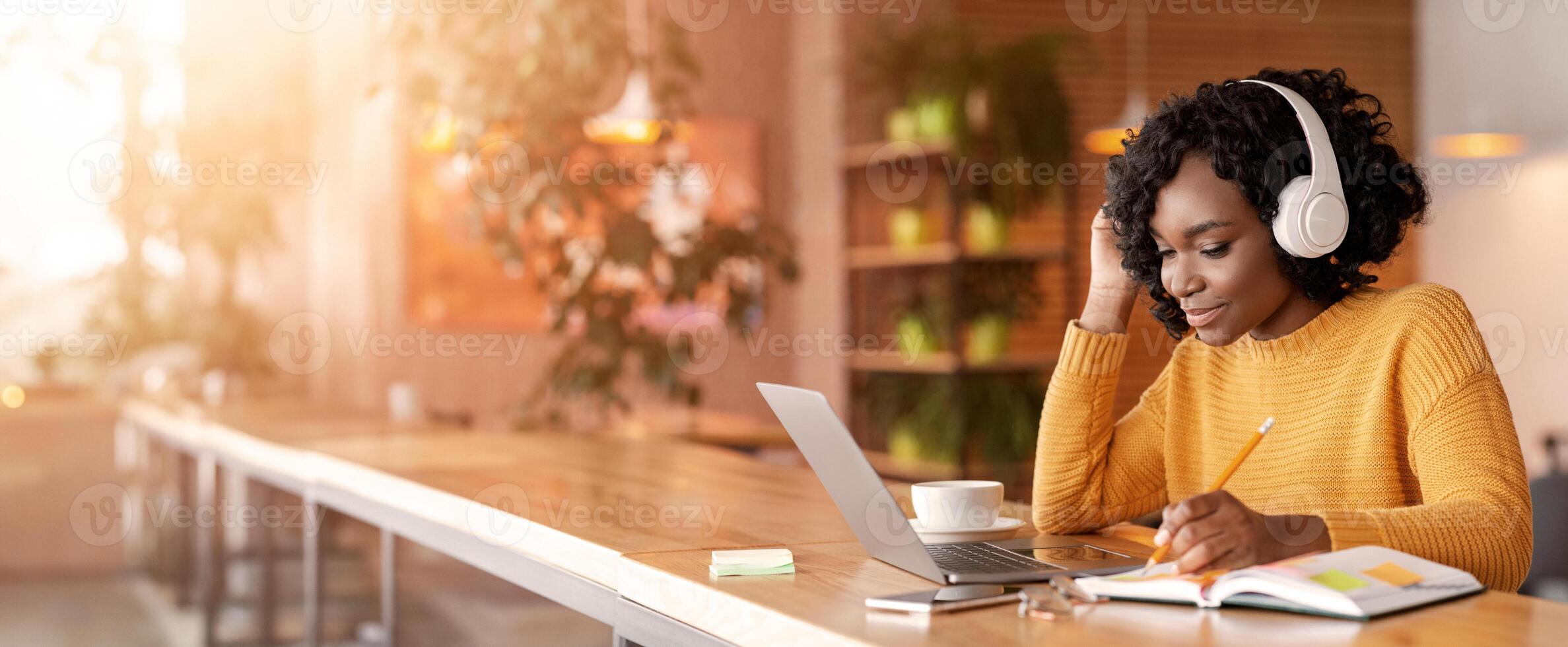 Smiling black girl with wireless headset studying online, using laptop at cafe, taking notes, copy space photo