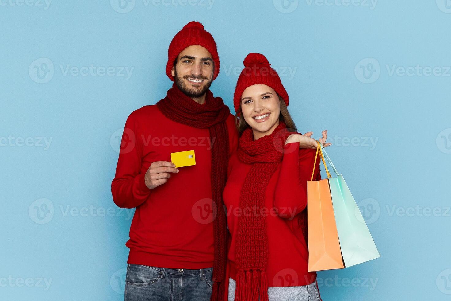 A young couple dressed in matching red winter clothing happily poses against a blue background, showcasing their shopping bags and a credit card, celebrating their recent purchases. photo