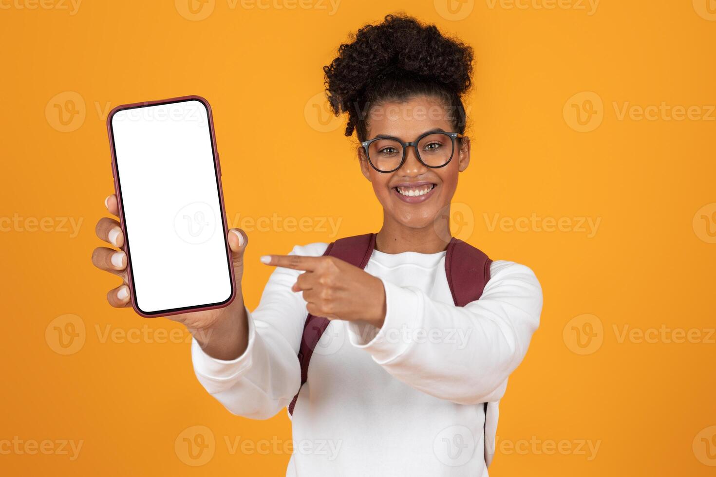 A cheerful young woman with curly hair holds a smartphone in front of her, pointing at the blank screen. The bright orange background adds energy to the scene, creating a lively atmosphere. photo