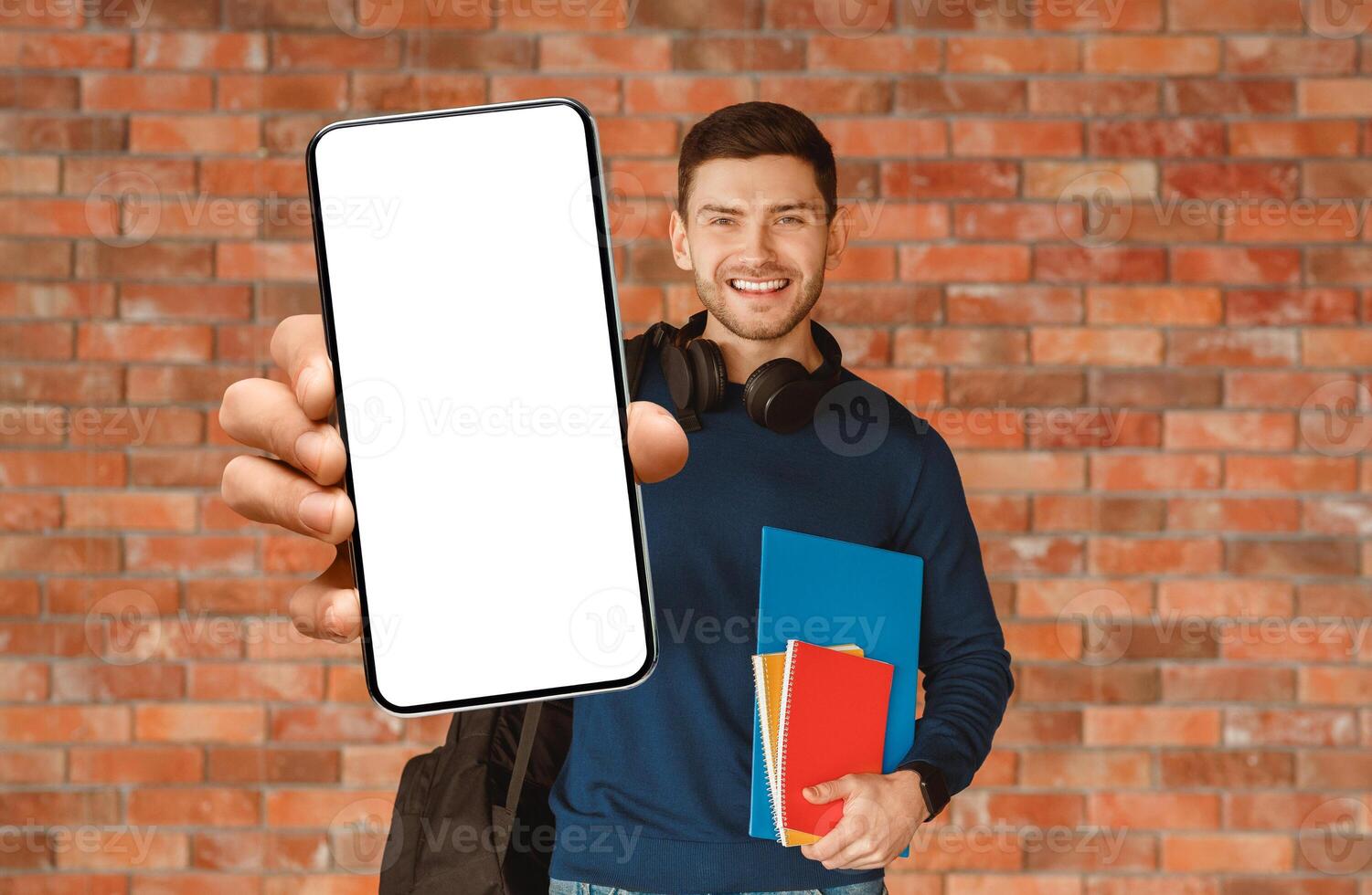 Modern Student. Happy Guy With Backpack Holding Smartphone And Notebooks Smiling To Camera Standing Over Brick Wall Background Indoor photo