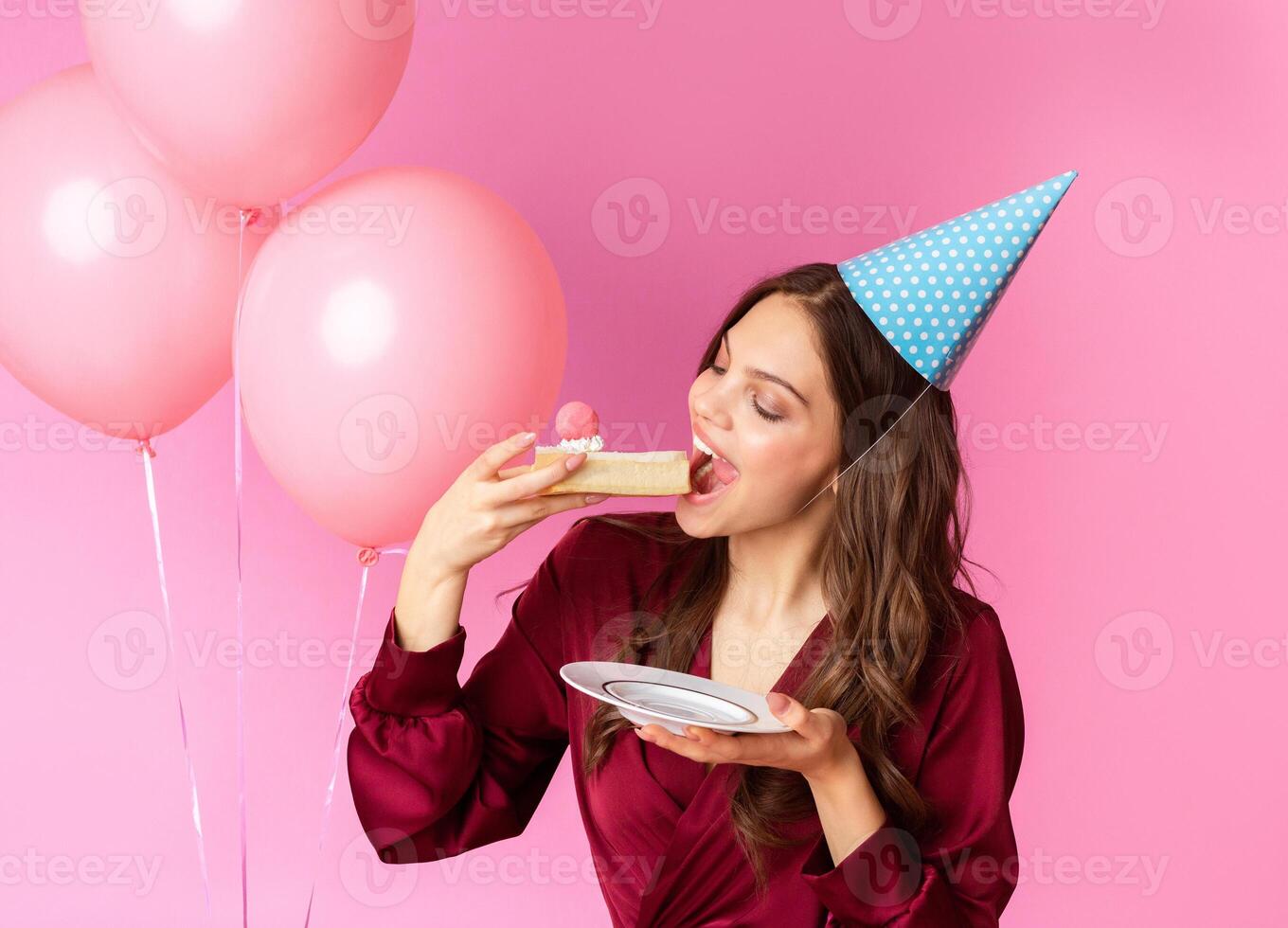 Birthday Lady Concept. Happy Young Woman In Party Hat Eating Piece Of B'Day Cake, Posing Over Colorful Background With Rosy Balloons photo