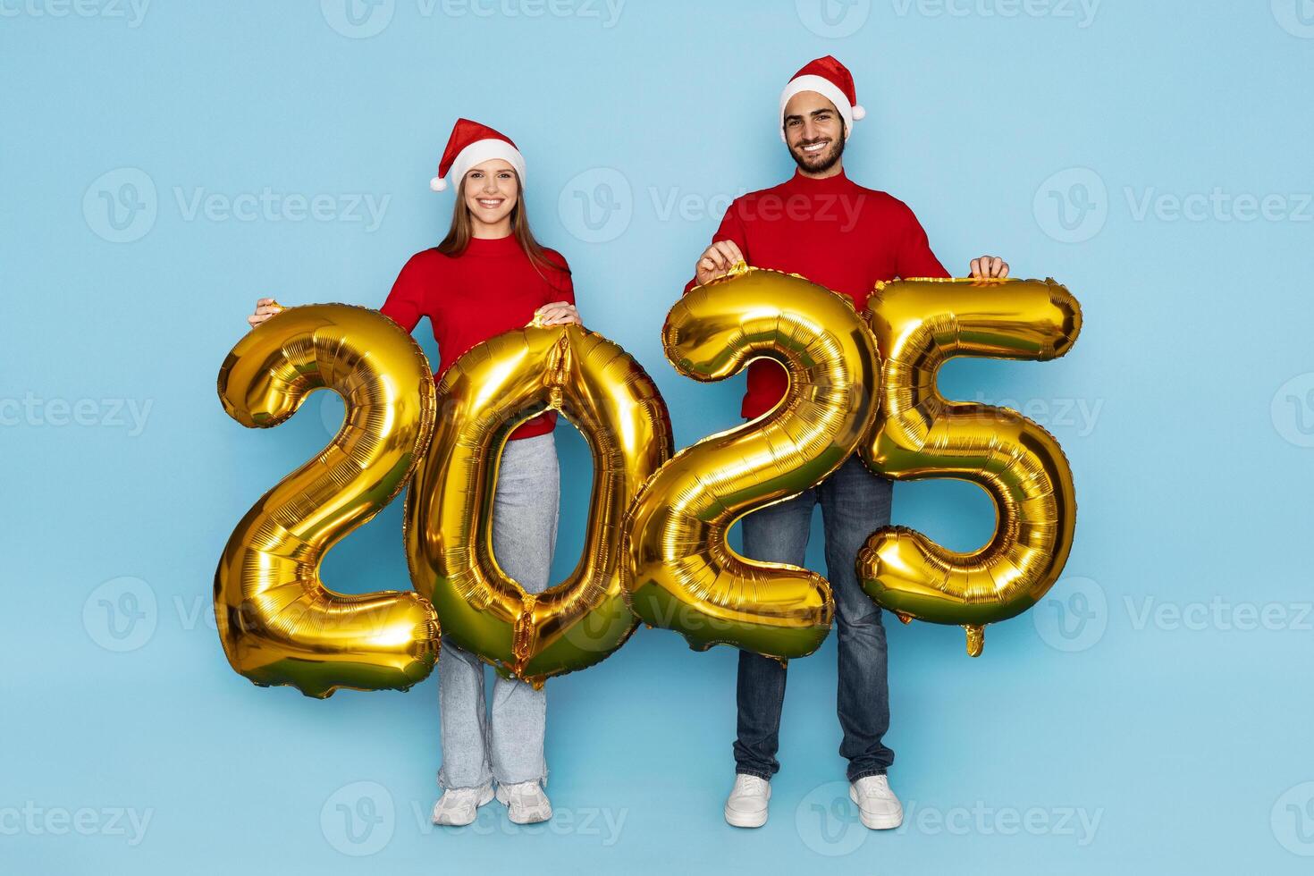 A man and woman in matching red sweaters and Santa hats cheerfully hold large golden balloons showing the year 2025, set against a bright blue background that enhances the festive atmosphere. photo