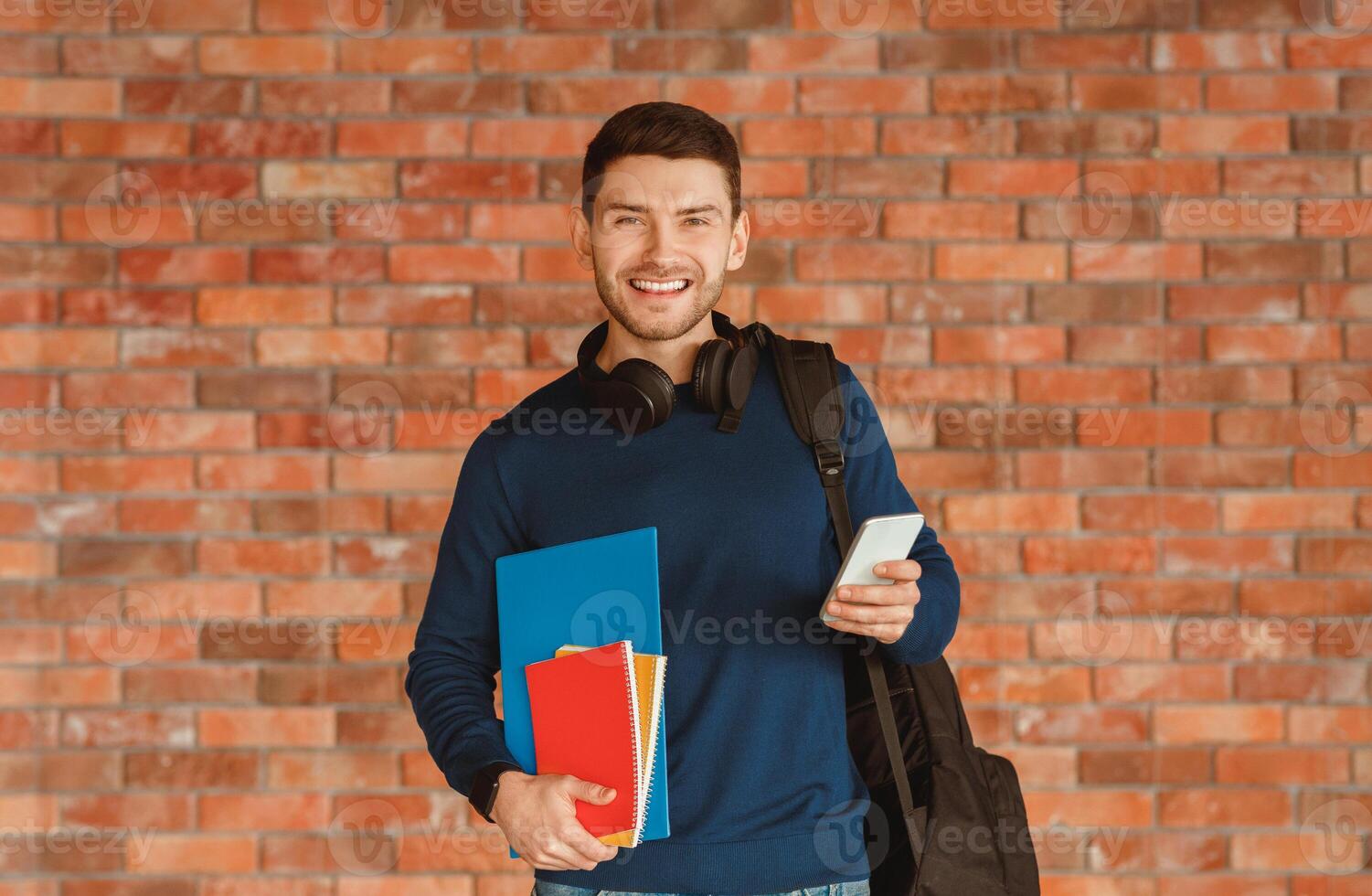 Modern Student. Happy Guy With Backpack Holding Smartphone And Notebooks Smiling To Camera Standing Over Brick Wall Background Indoor photo