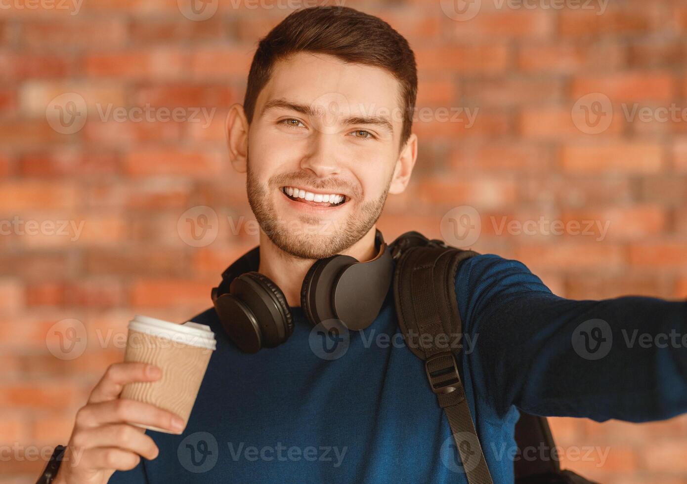 Positive Guy Making Selfie Smiling To Camera And Holding Paper Coffee Cup Standing Over Brick Wall Indoor photo