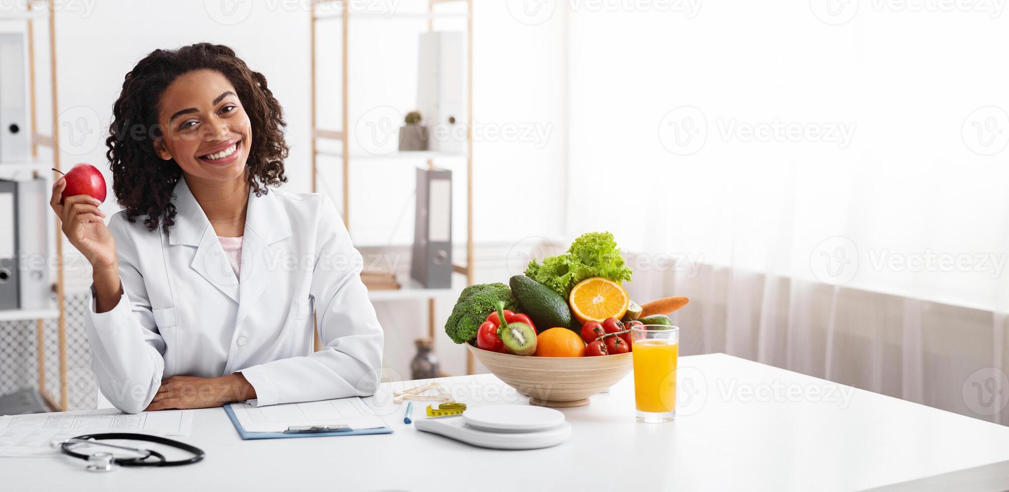 Diet while lockdown. Pretty black lady dietologist holding apple in her hand and smiling, recommending fresh fruits, posing at clinic, copy space photo