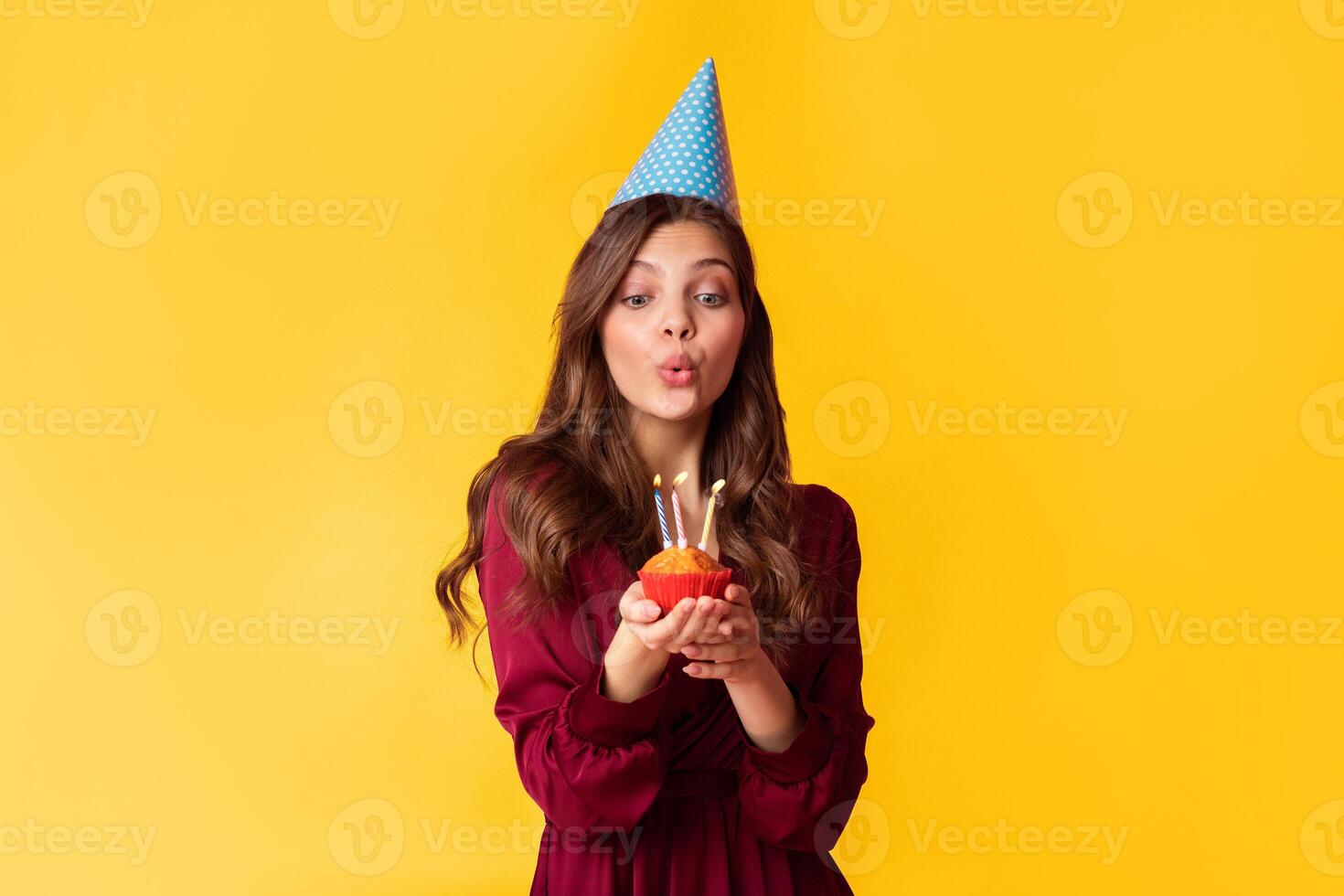 Beautiful lady in party hat blowing candles on birthday cupcake, making wish, celebrating B'Day, posing over colorful studio background with copy space photo