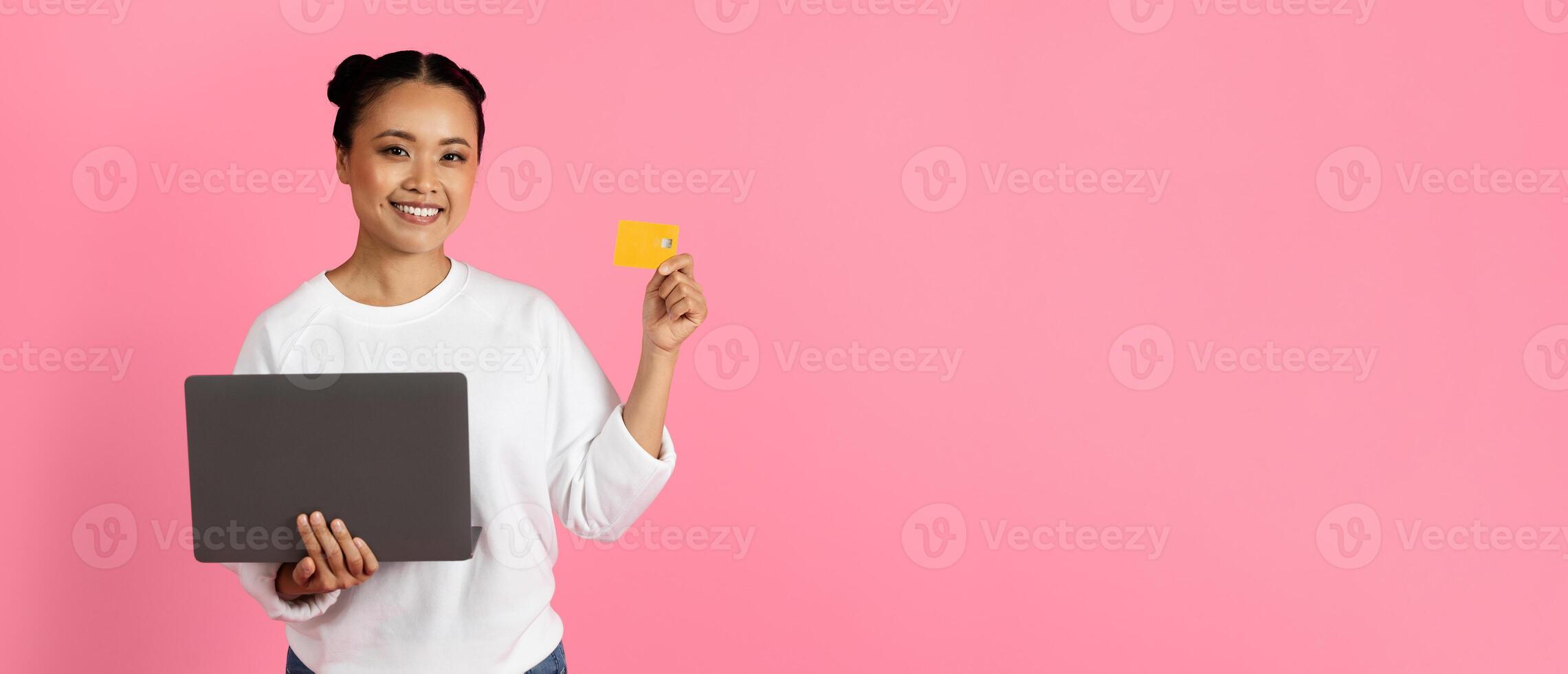 A young woman smiles brightly while holding a laptop in one hand and a credit card in the other, set against a vibrant pink background, conveying a sense of digital empowerment, copy space photo