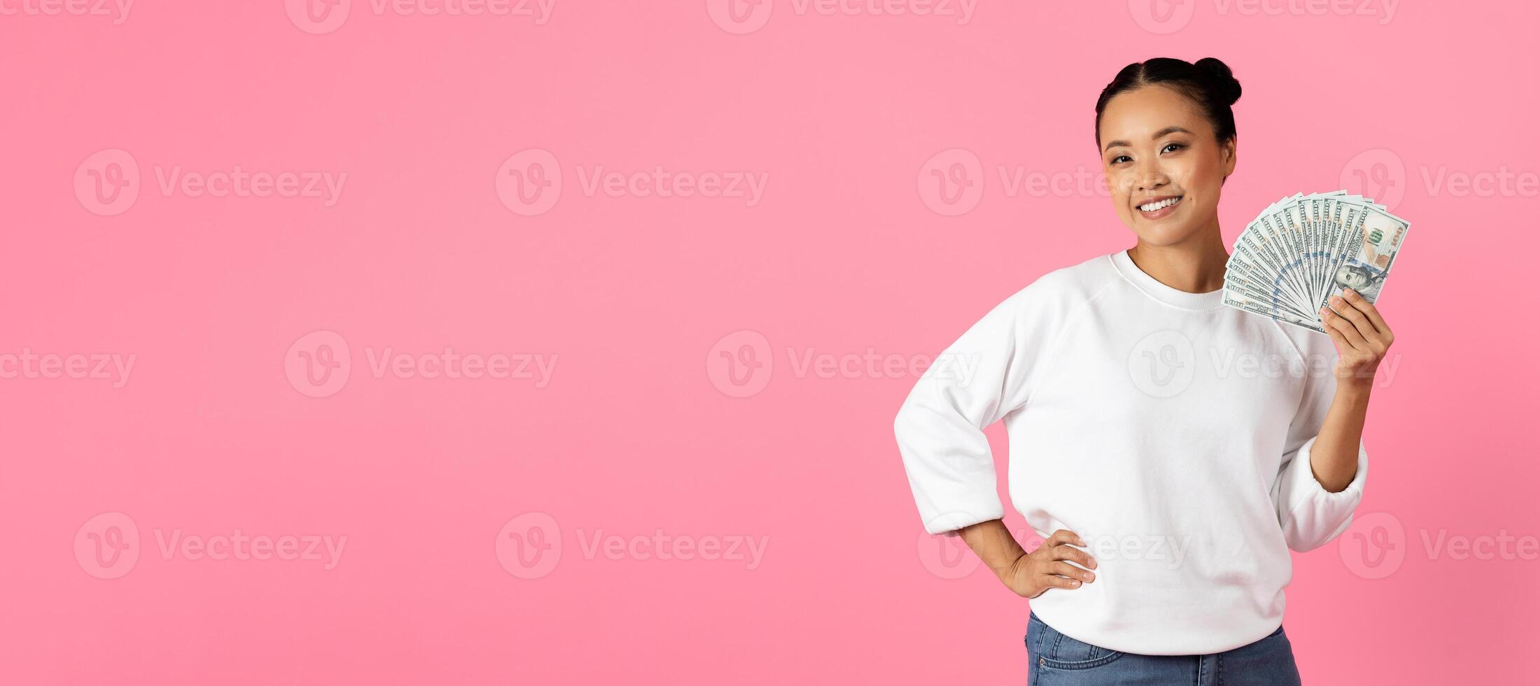 A woman stands confidently with a smile, showing off a fan of cash in her hand. The vibrant pink background emphasizes her joyful expression and casual outfit, copy space photo