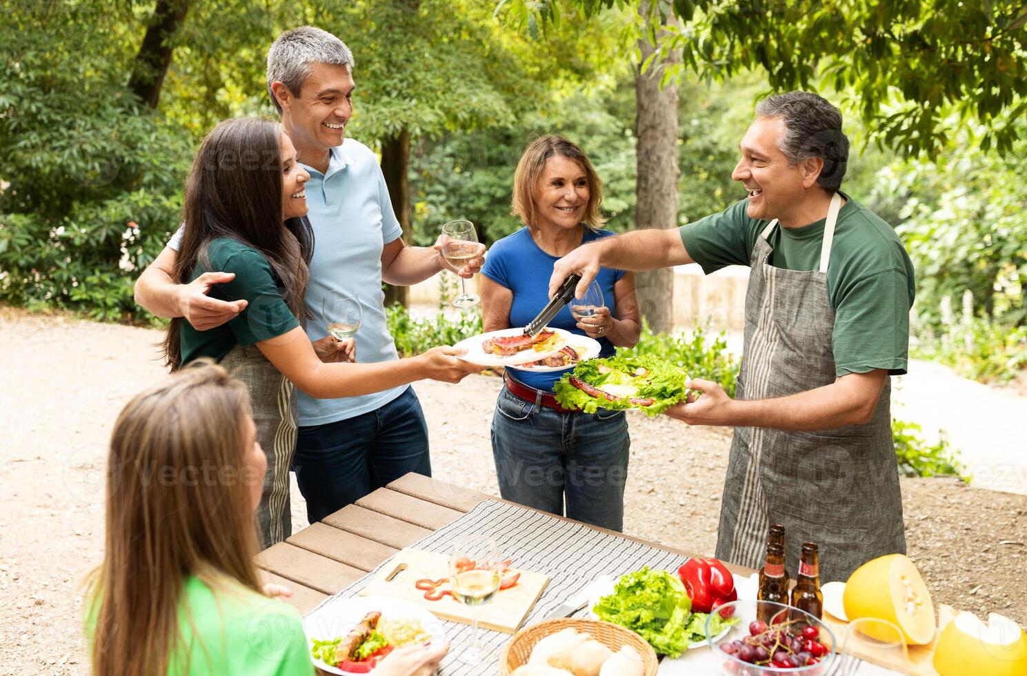 A group of friends and family shares a joyful moment at a picnic table, enjoying a barbecue meal while surrounded by greenery on a sunny day. Laughter and conversation fill the air. photo