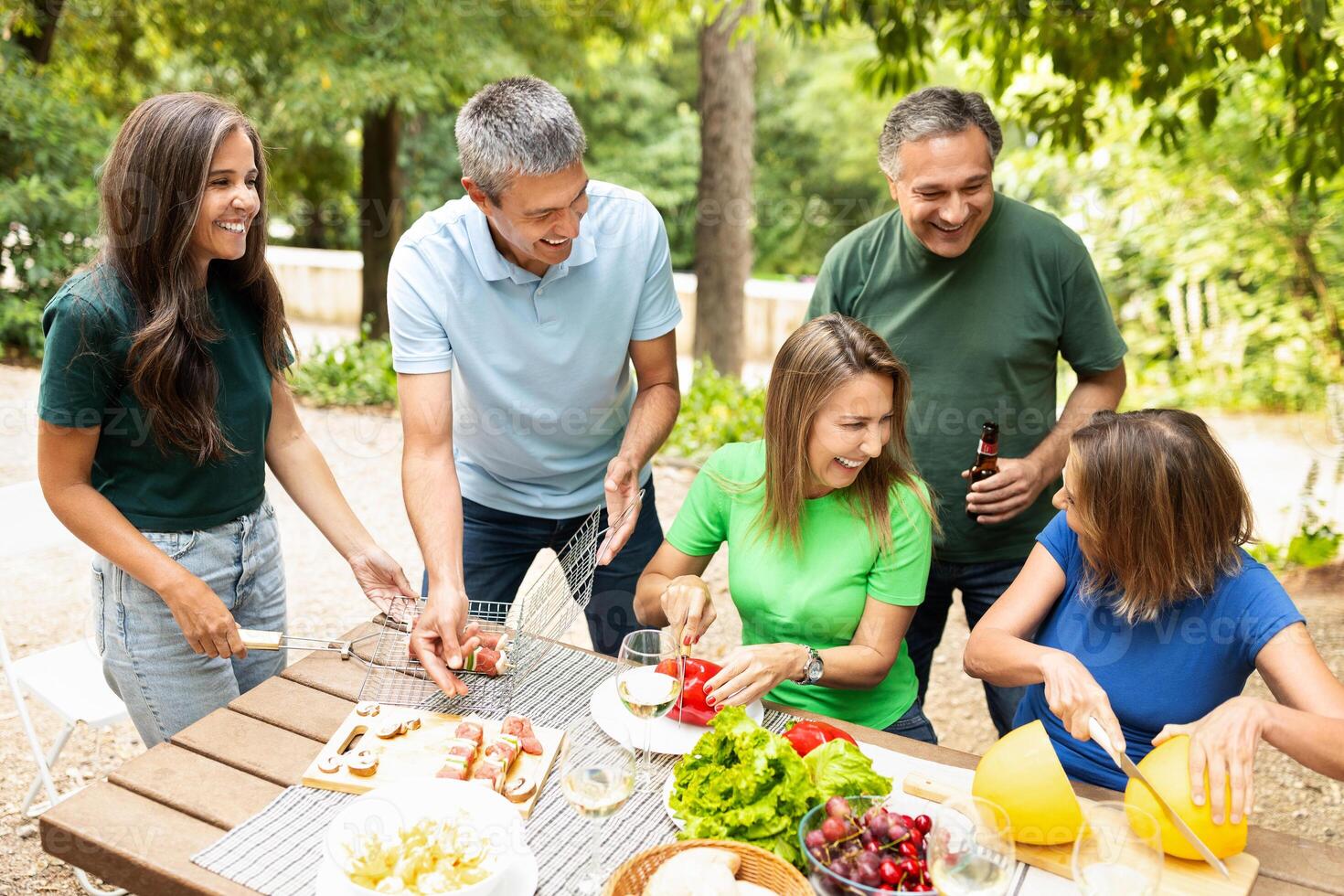 A lively group of friends shares laughter and delicious food while grilling in a green park. They prepare fresh vegetables and enjoy refreshing drinks in the sunny atmosphere. photo