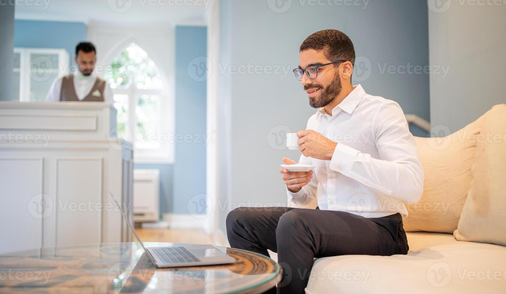 A man in a white shirt sips coffee and smiles while working on his laptop in a cozy lounge. A staff member can be seen in the background providing service, creating a relaxed atmosphere. photo