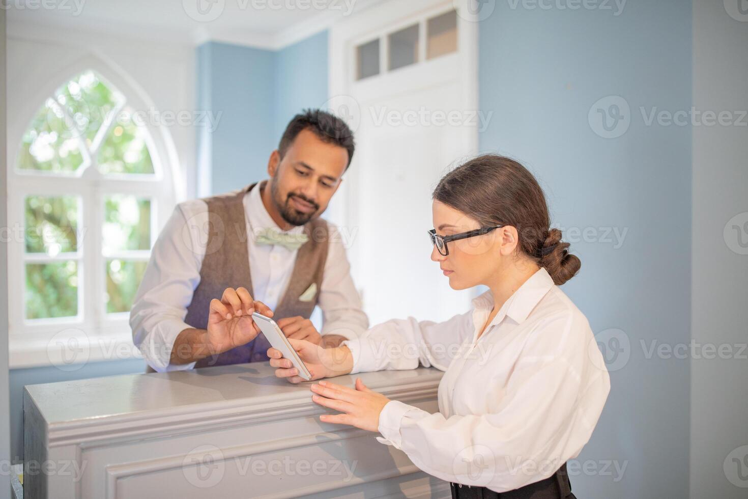 A man in formal attire helps a woman at a reception desk. She is using her smartphone while they discuss details in a well-lit, stylish environment. photo