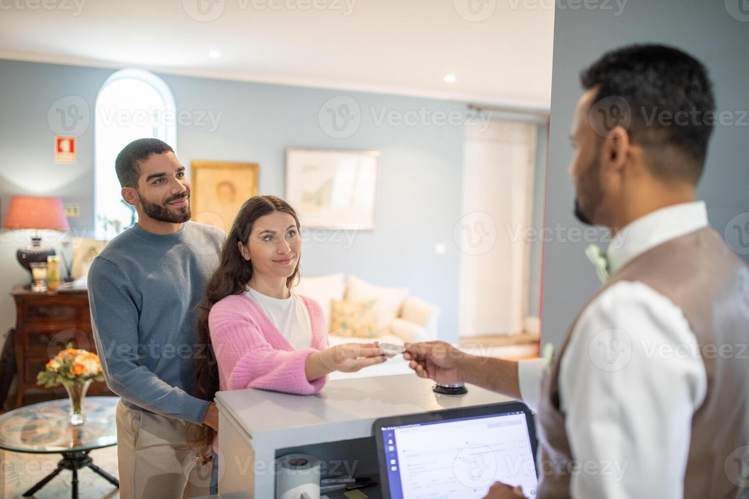 A couple hands over their identification to a hotel receptionist at a welcoming front desk, surrounded by a cozy lobby adorned with flowers and soft lighting. photo