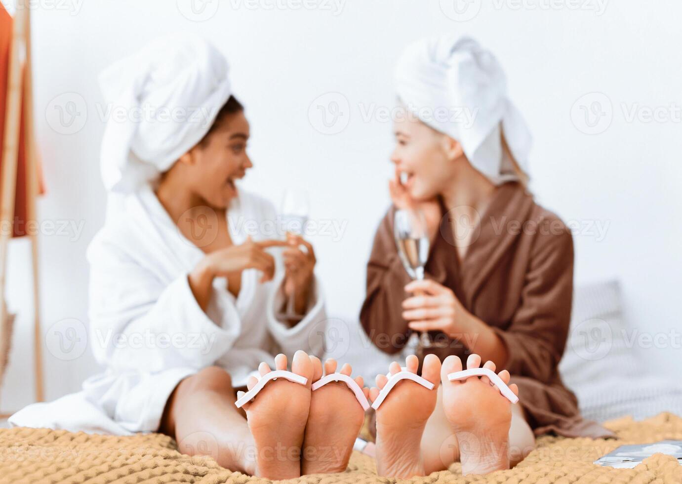 Multiracial girlfriends having spa day at home, drinking champagne, making pedicure, sharing secrets photo