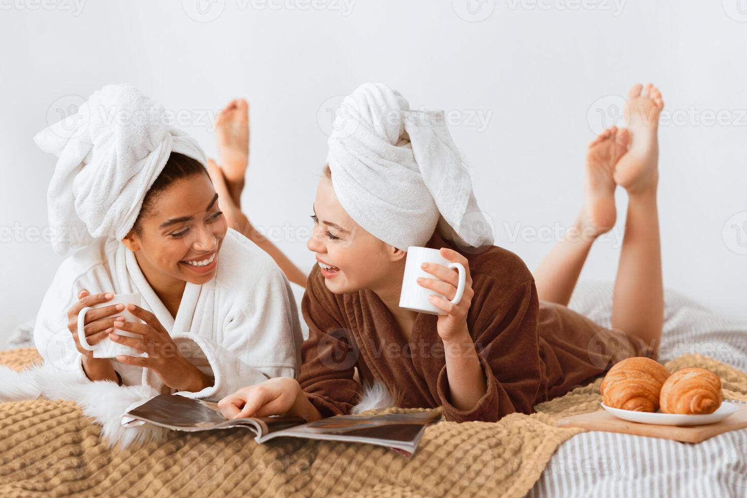 Smiling beautiful multiracial women in bathrobes and towels on heads drinking coffee, reading magazines together while laying on bed at home photo