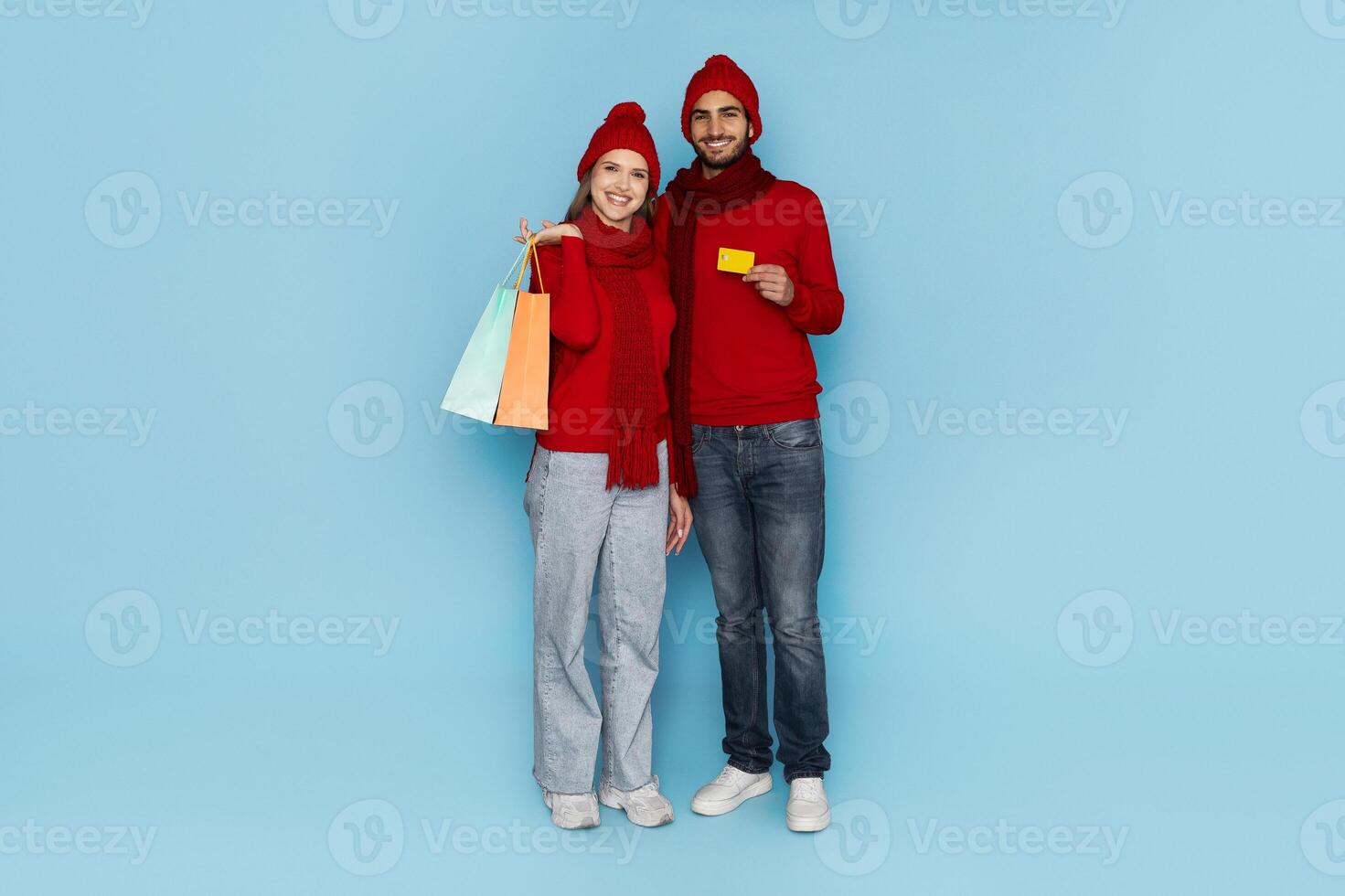 A happy couple poses together, showcasing colorful shopping bags and a gift card. They wear matching red sweaters and hats, smiling joyfully in a bright blue environment. photo