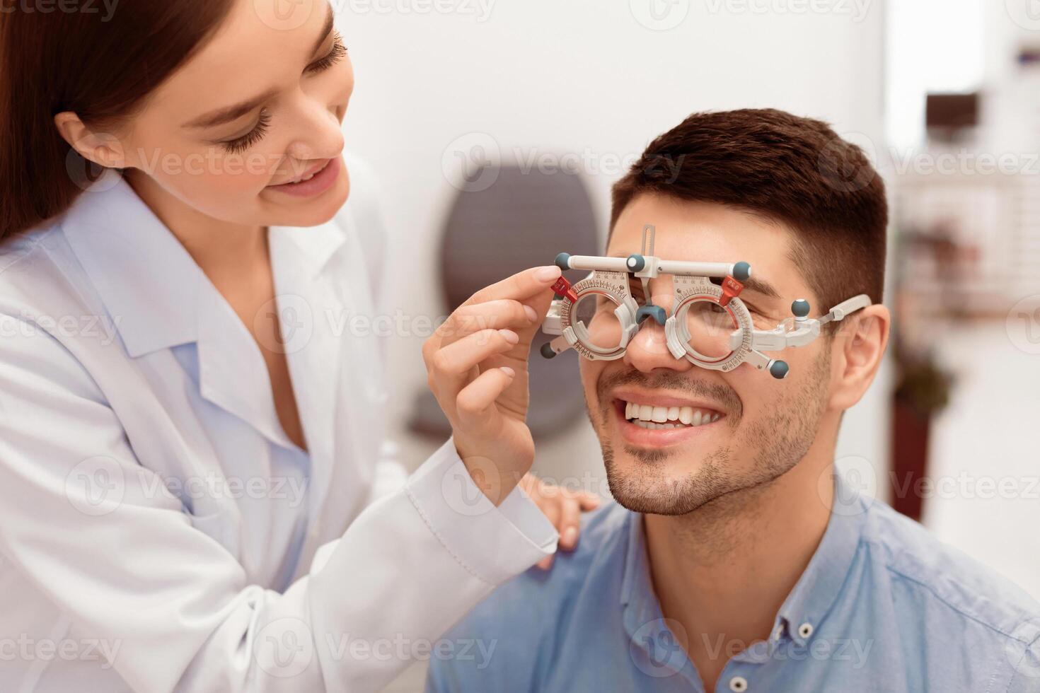Eye Test Concept. Beautiful lady specialist checking vision of male client with trial frame, touching tool photo