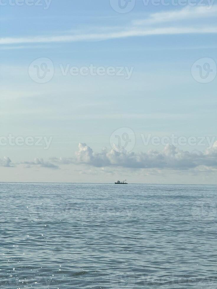 A small boat is seen far on the horizon, floating on a calm ocean under a bright and clear sky with a few scattered clouds. Peaceful maritime scene photo