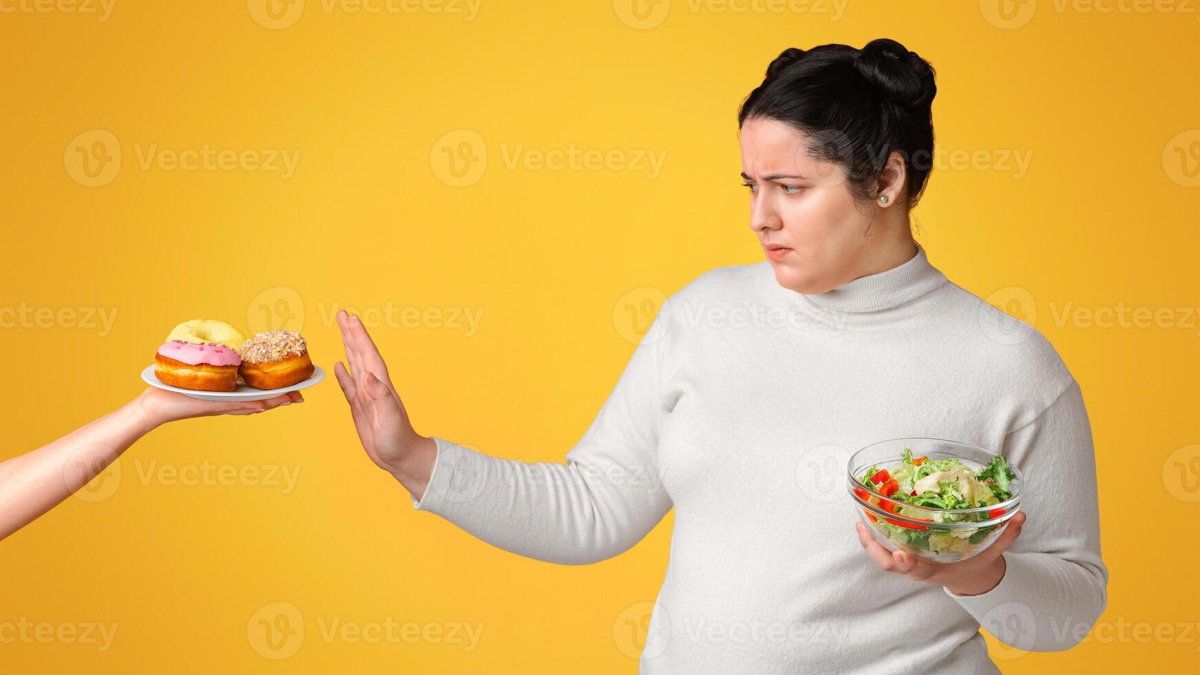 Healthy life concept. Young plump girl refusing junk food, holding bowl with vegetable salad, choosing greens over donuts, yellow background, panorama photo