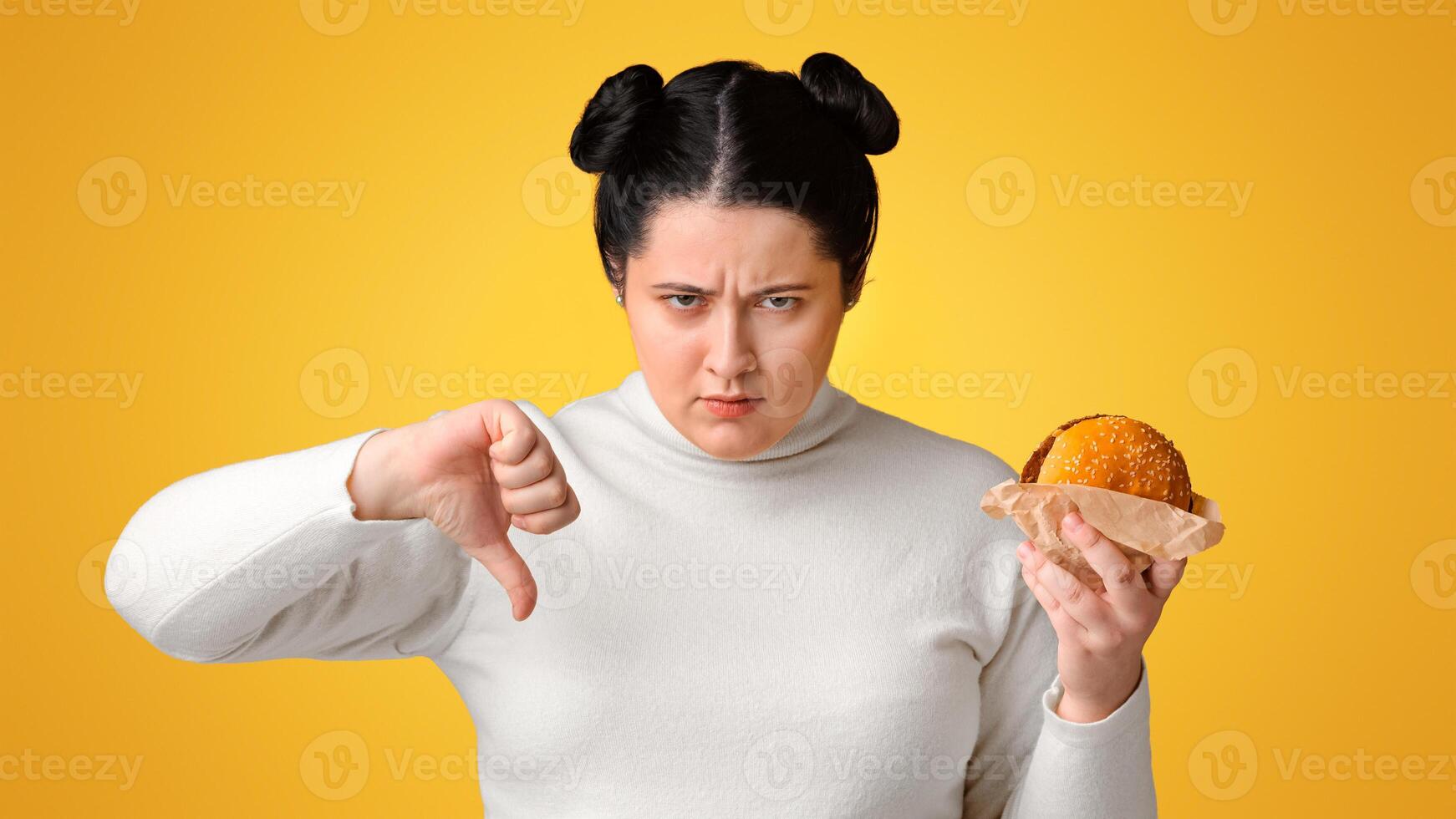 Stop Eating Fast Food. Annoyed overweight girl holding hamburger and showing thumb down, rejecting unhealthy meal, yellow background with free space photo
