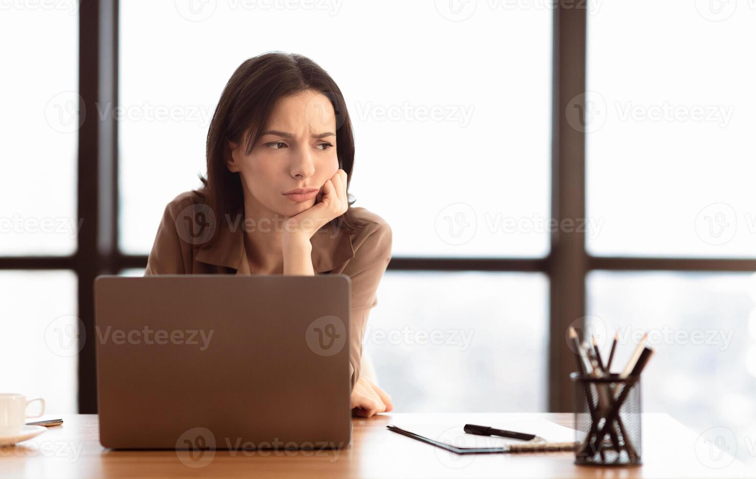 Boredom. Portrait of sad young female manager using laptop sitting at table in modern office, looking away, copyspace photo