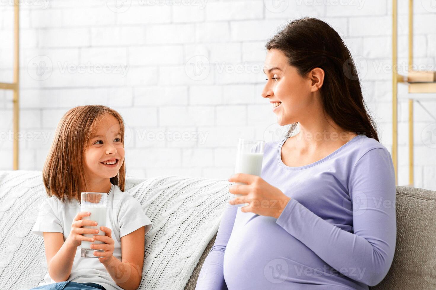 Pregnant loving mother drink milk with her smiling daughter, sitting on sofa at living room photo