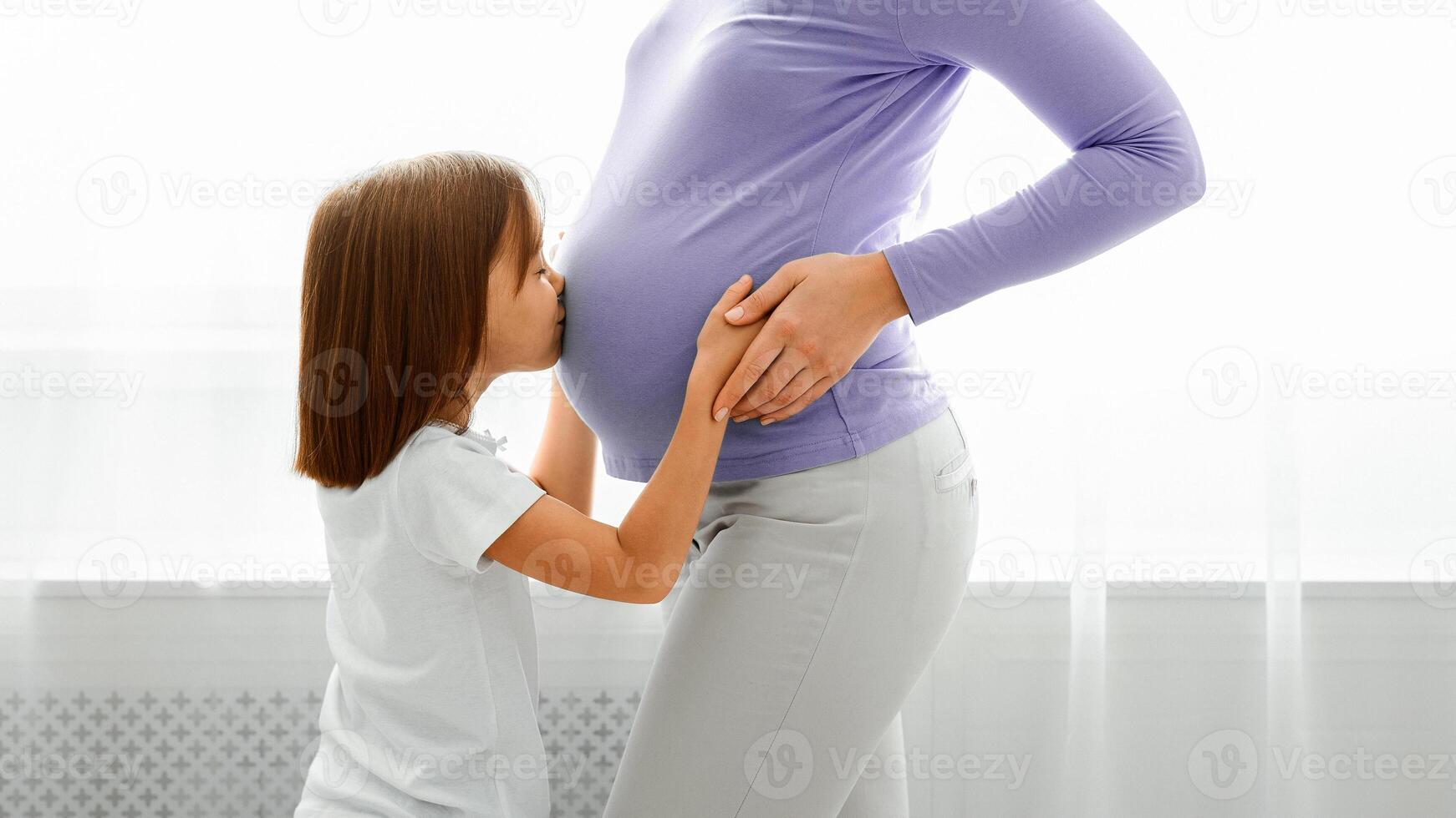 Close up of little daughter kissing her pregnant mom tummy, white background photo