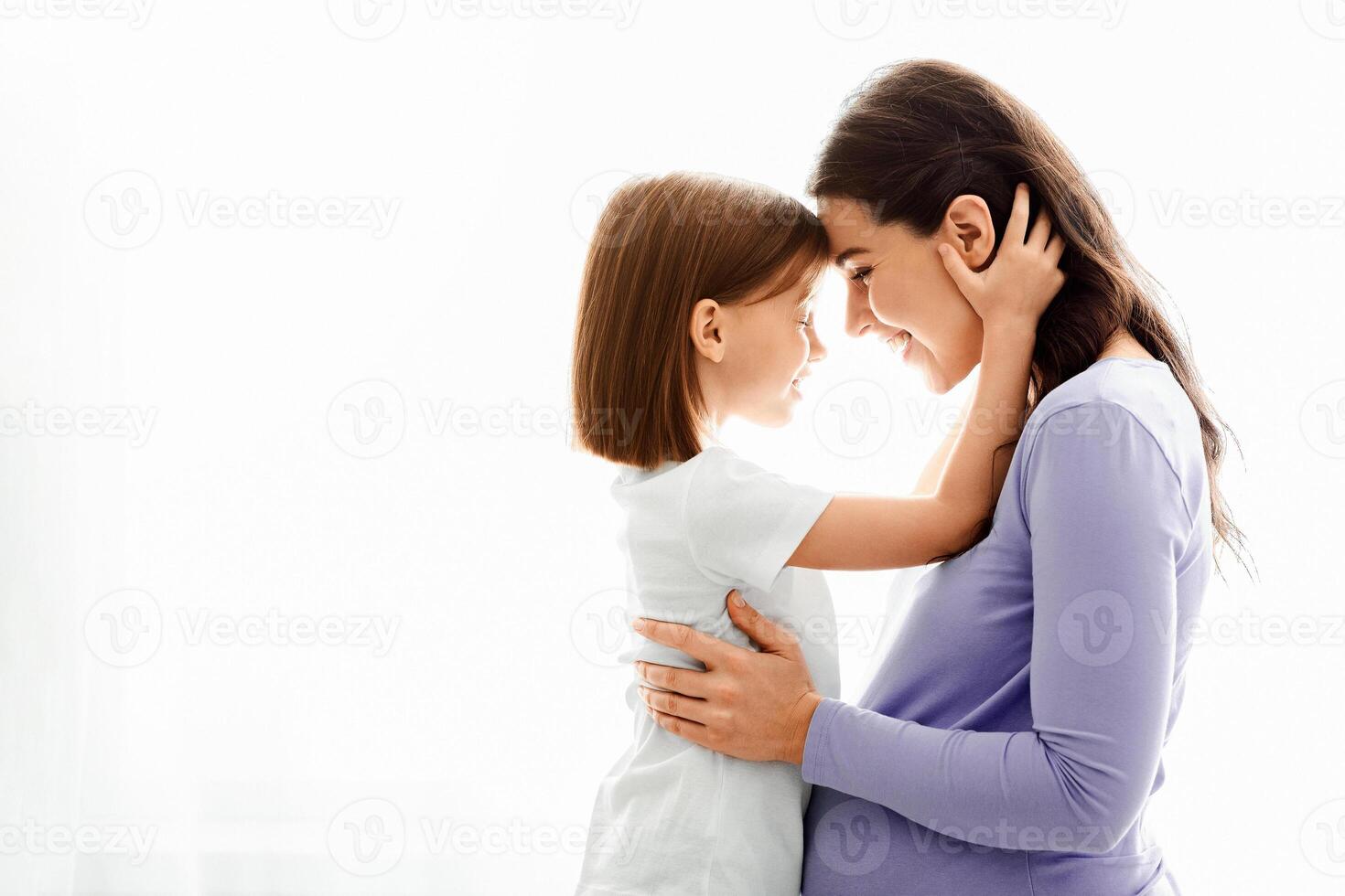 Loving little daughter embracing her expecting young mother, looking at her eyes, side view, window background photo