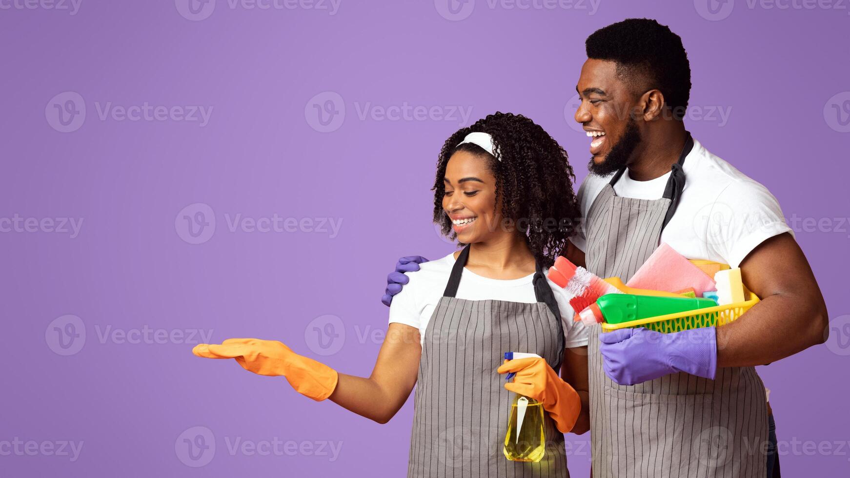 Best Detergent. Couple of professional cleaners holding bucket with cleaning supplies and demonstrating invisible object on open palm, pink background photo