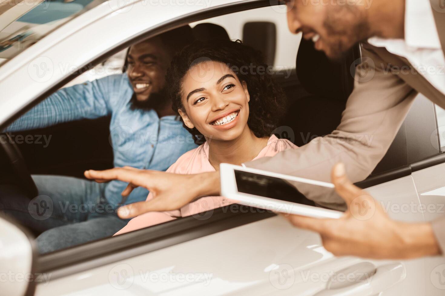 Happy Black Couple Buying New Car Testing And Talking With Salesman Sitting In Auto Dealership Showroom. Family Choosing Automobile. photo