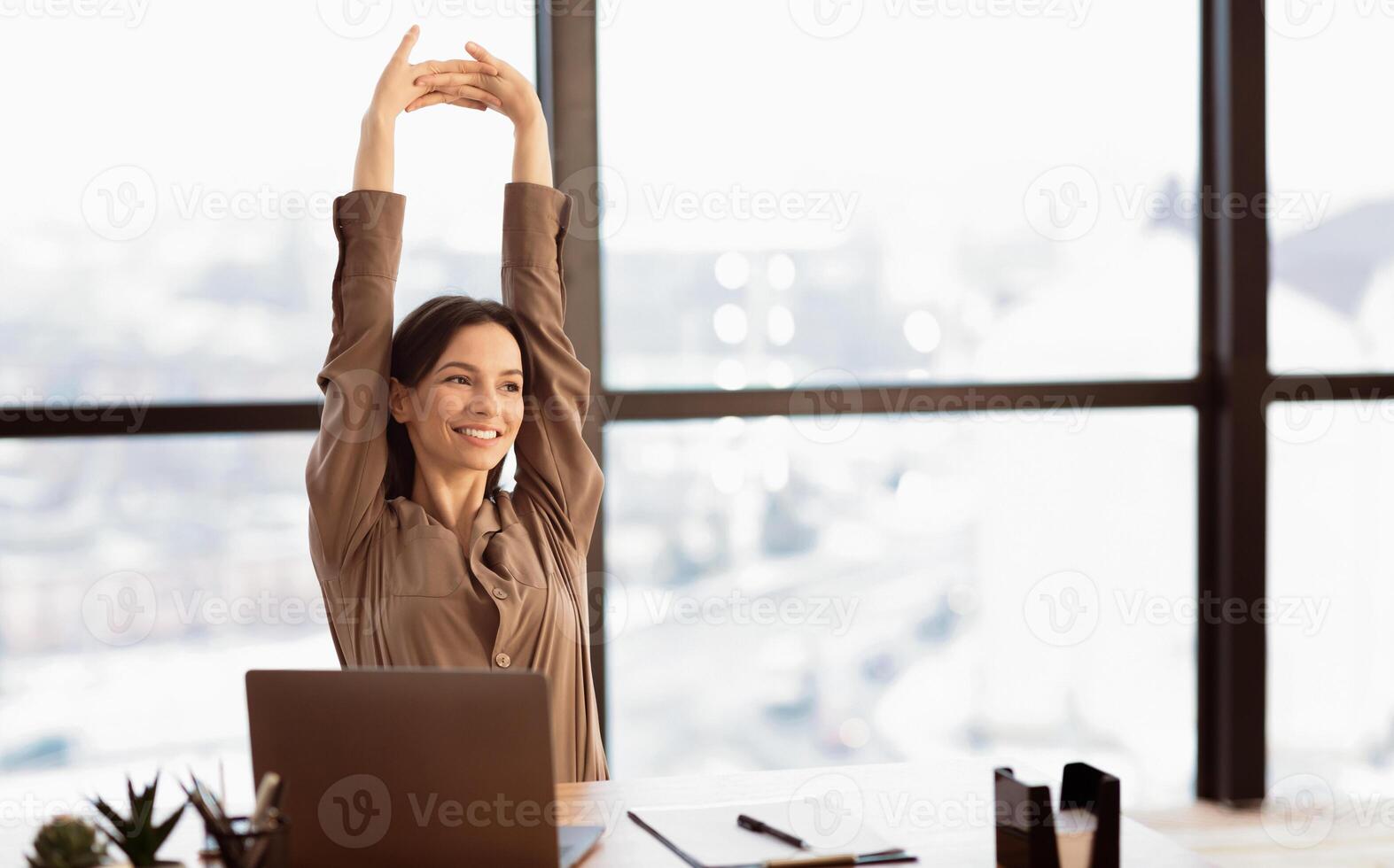 Stretching At Workplace. Happy beautiful young woman sitting at desk in modern office, looking away, copyspace photo