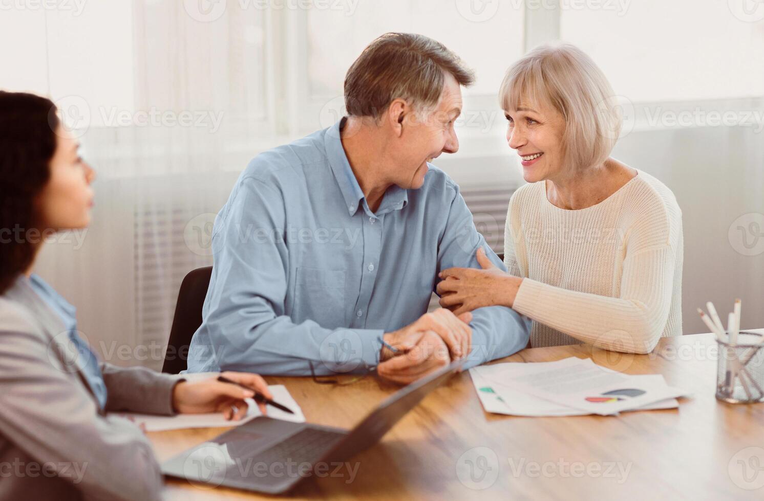 Best Offer. Happy mature couple looking at each other, sitting at the desk at office with financial consultant photo