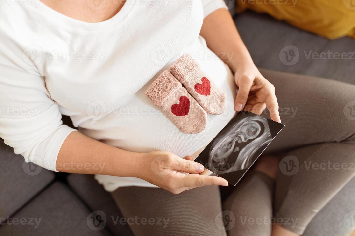 A pregnant woman comfortably sits on a couch, holding an ultrasound image and baby booties adorned with hearts, cherishing the anticipation of her child arrival. photo