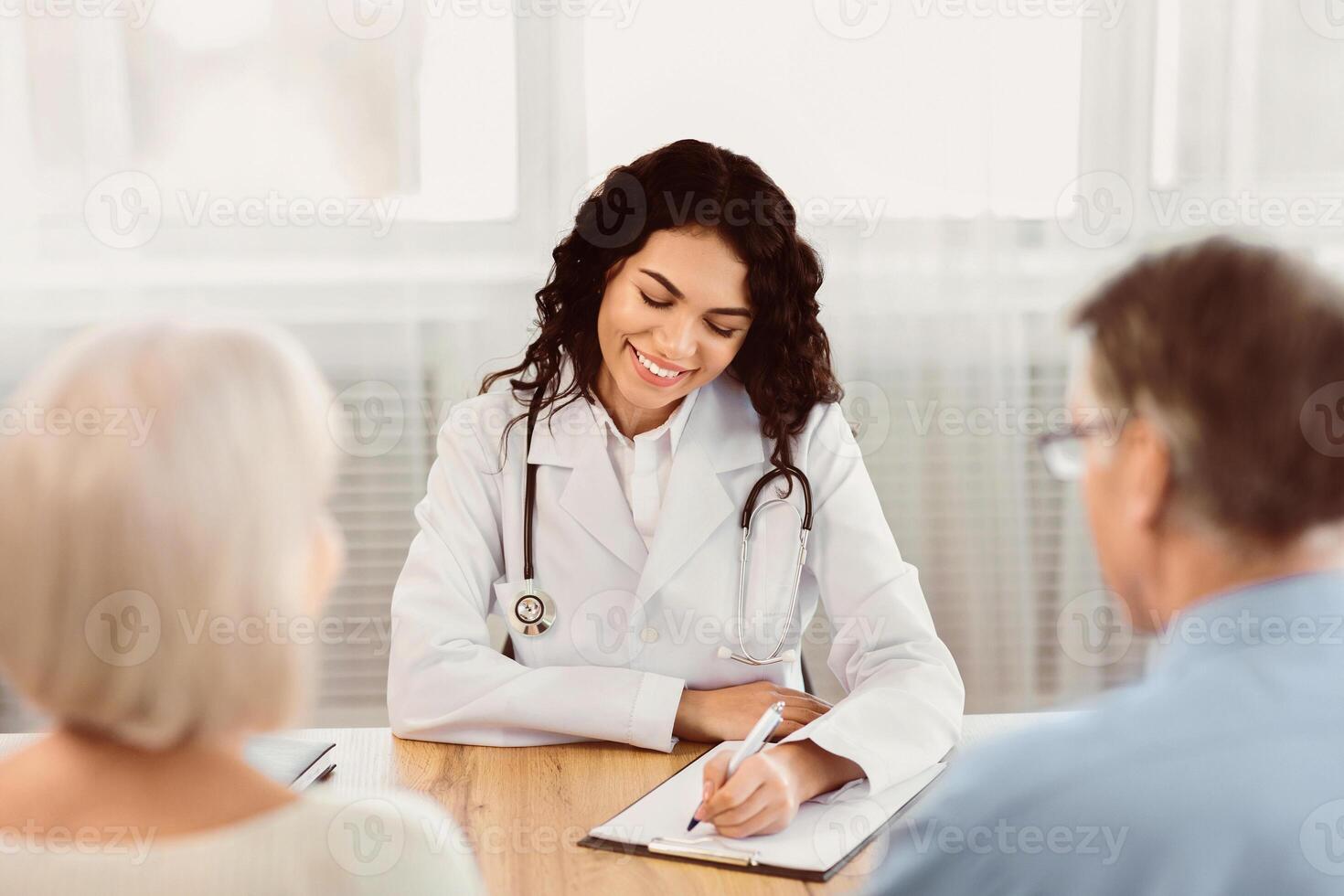 Consultation Concept. Happy latina doctor taking notes about symptoms, filling out medical prescription to senior couple photo
