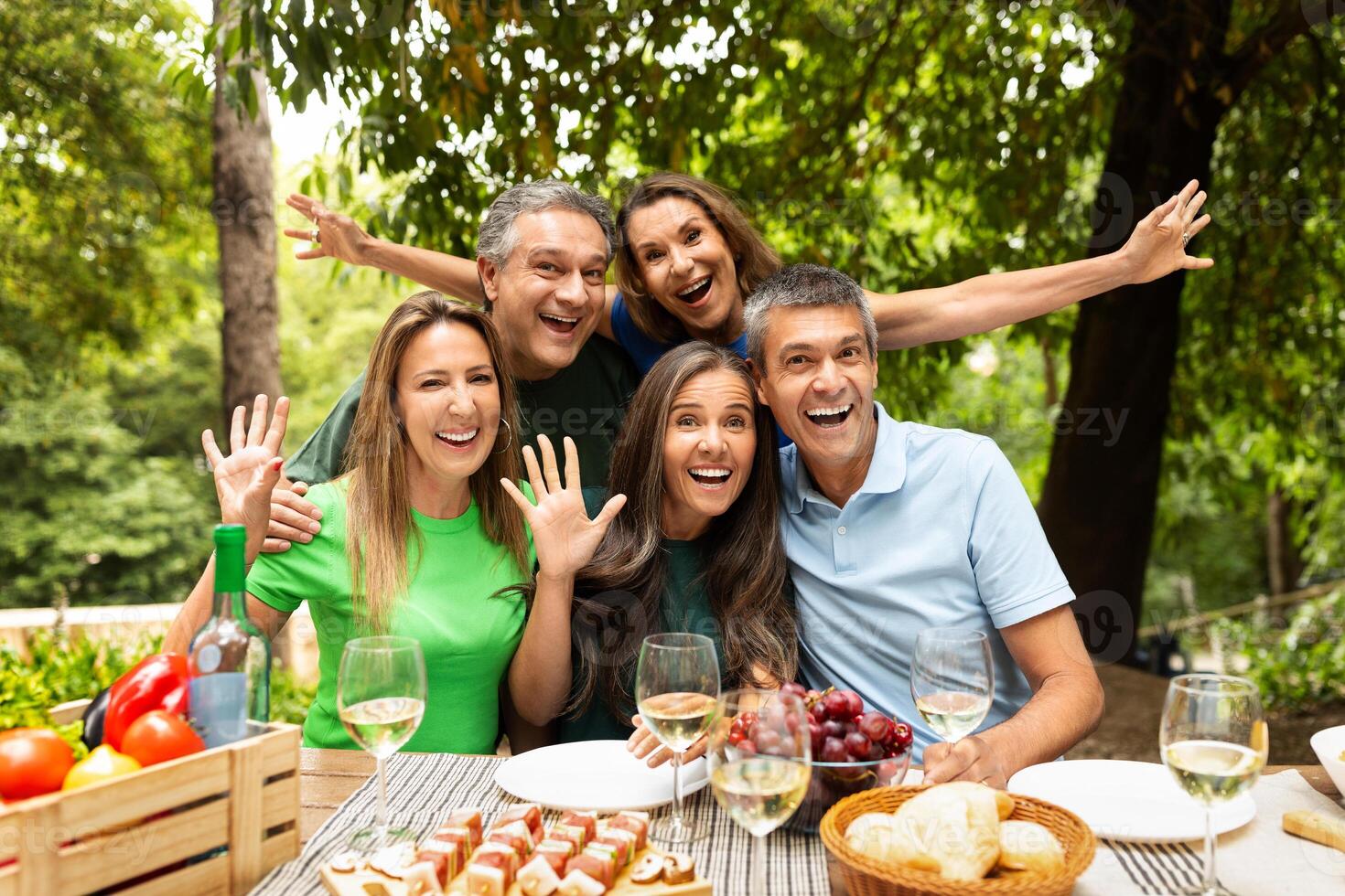 A group of five friends joyfully poses for a picture during a lively outdoor picnic, surrounded by delicious food, drinks, and greenery in the afternoon sunlight. photo