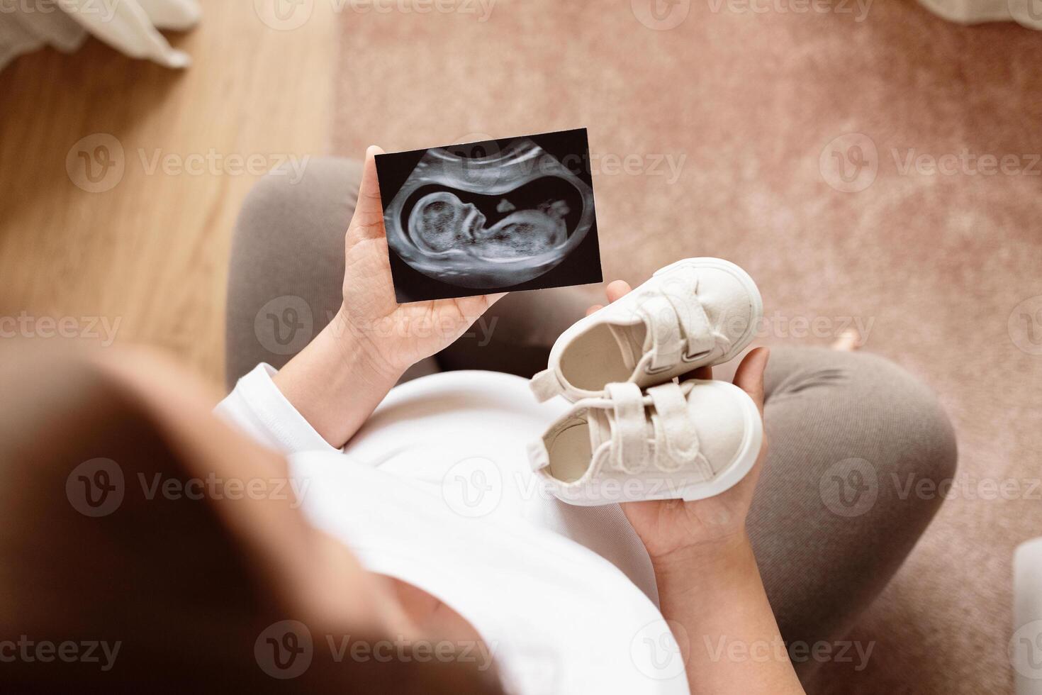 A pregnant woman is sitting comfortably, looking at an ultrasound image while holding a pair of baby shoes. The sense of anticipation and love fills the warm room. photo