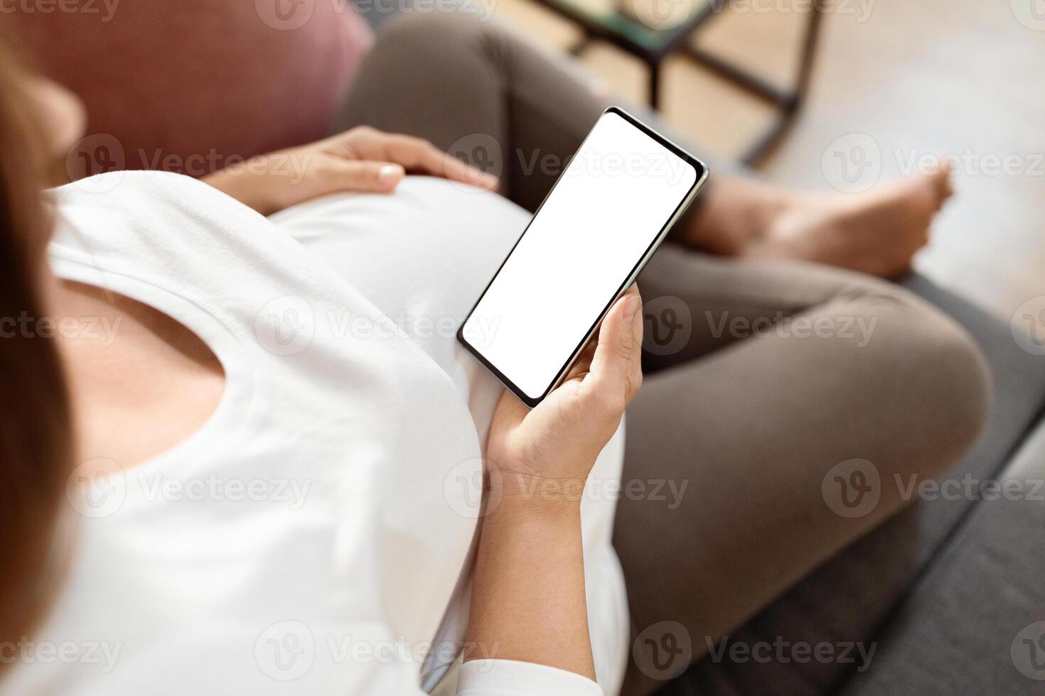 A pregnant woman is comfortably seated on a couch, holding her smartphone, looking relaxed. She is dressed casually, enjoying a peaceful moment at home in the afternoon. photo