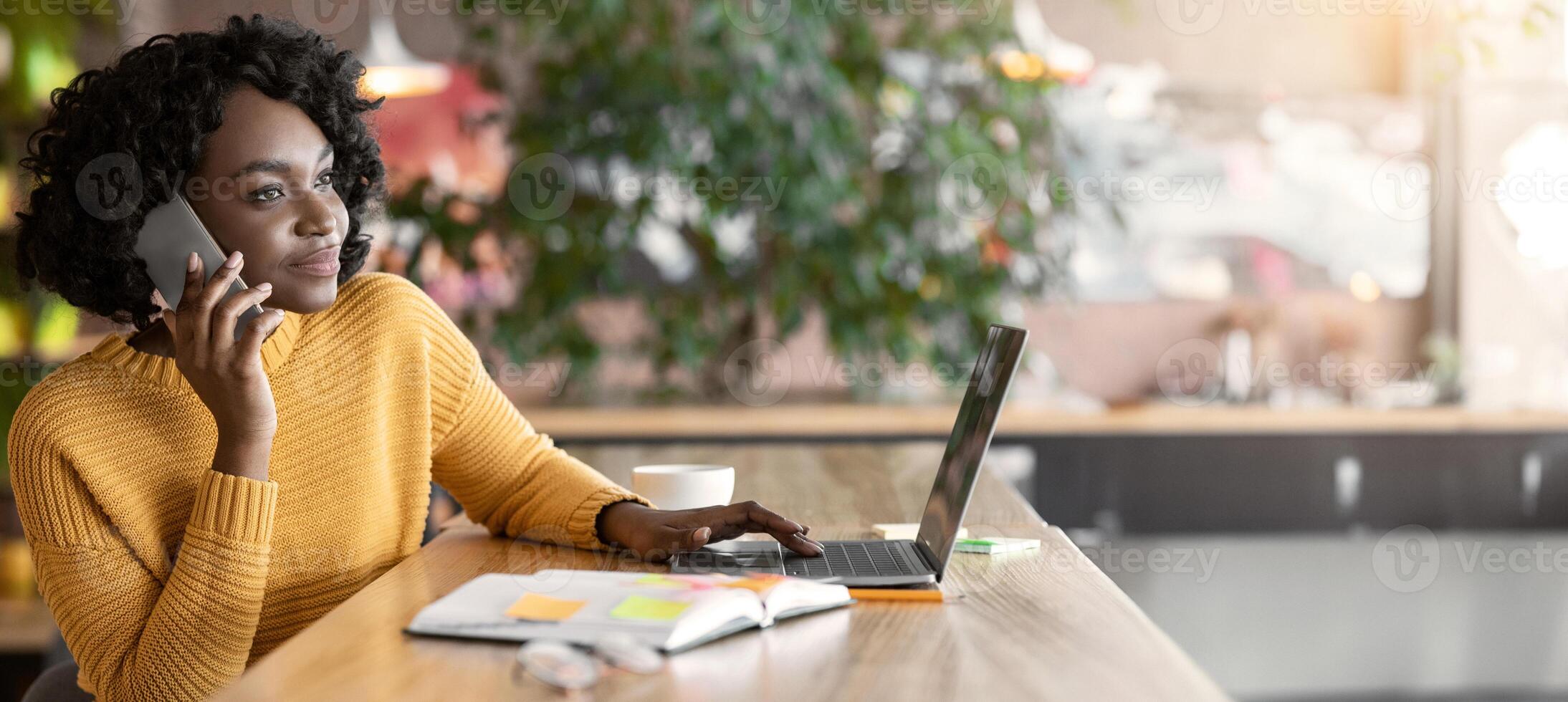 Young black businesswoman having conversation with clients on phone, working at cafe, using laptop, free space photo
