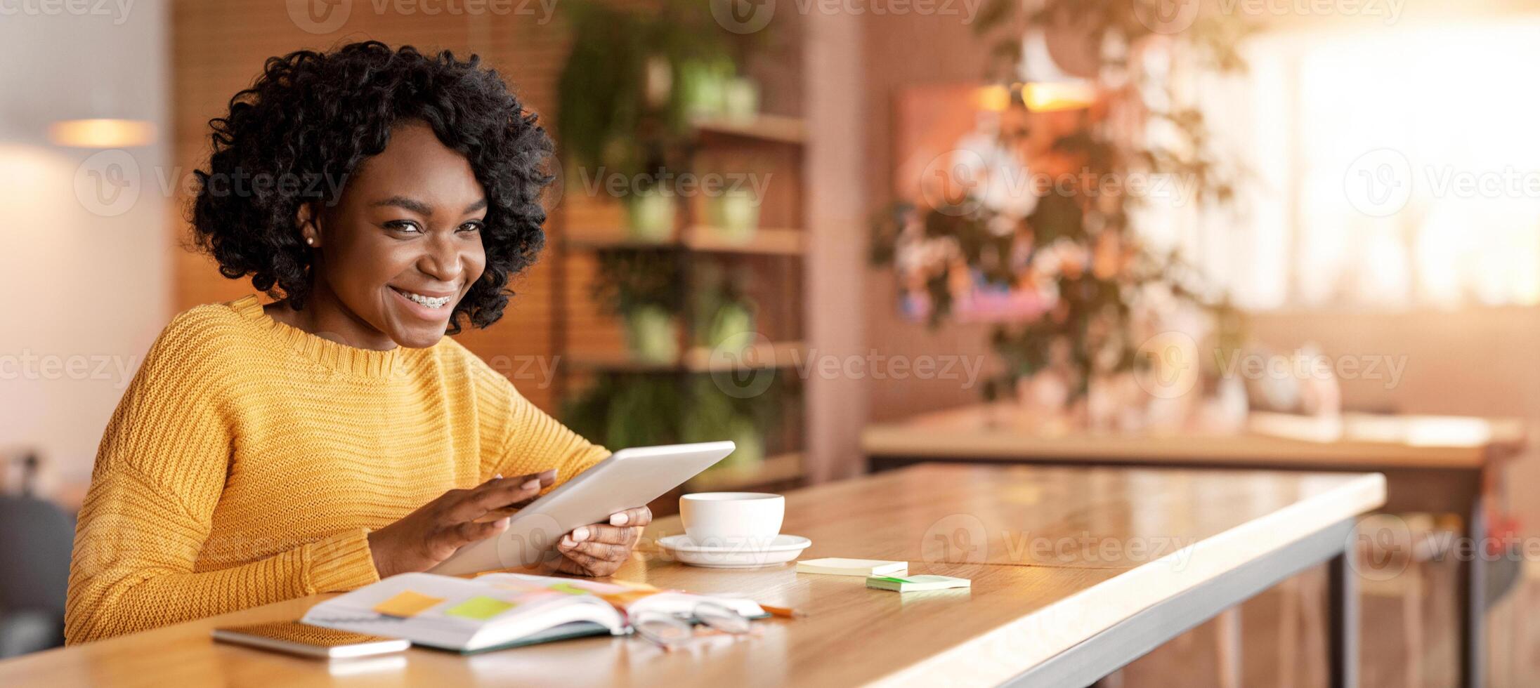 Cheerful african girl using social media on digital tablet while drinking coffee at cafe, copy space photo