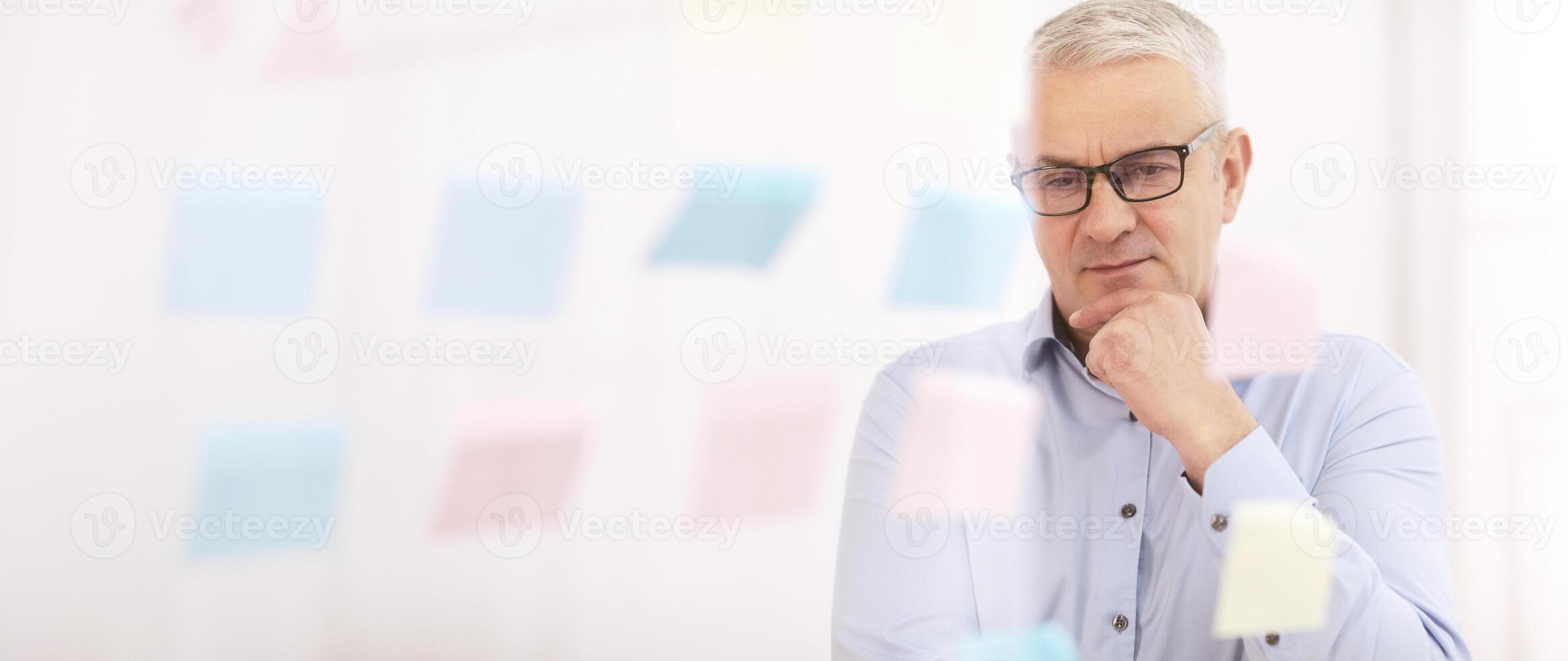 Brainstorming. Thoughtful mature businessman in specs looking at sticky notes on glass wall, touching chin, empty space photo