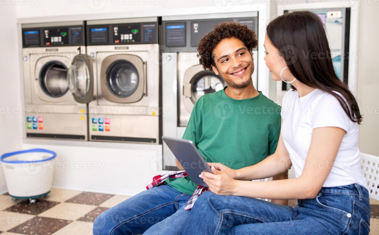 A young couple sits together in a laundromat, smiling and engaged with a tablet. Clean laundry machines fill the background, indicating a relaxed afternoon atmosphere. photo
