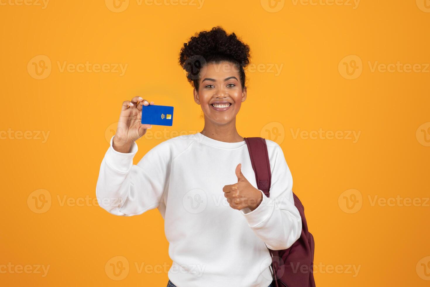 A cheerful young woman stands against a bright orange backdrop, showcasing her credit card with one hand while giving a thumbs-up with the other, expressing excitement and confidence. photo