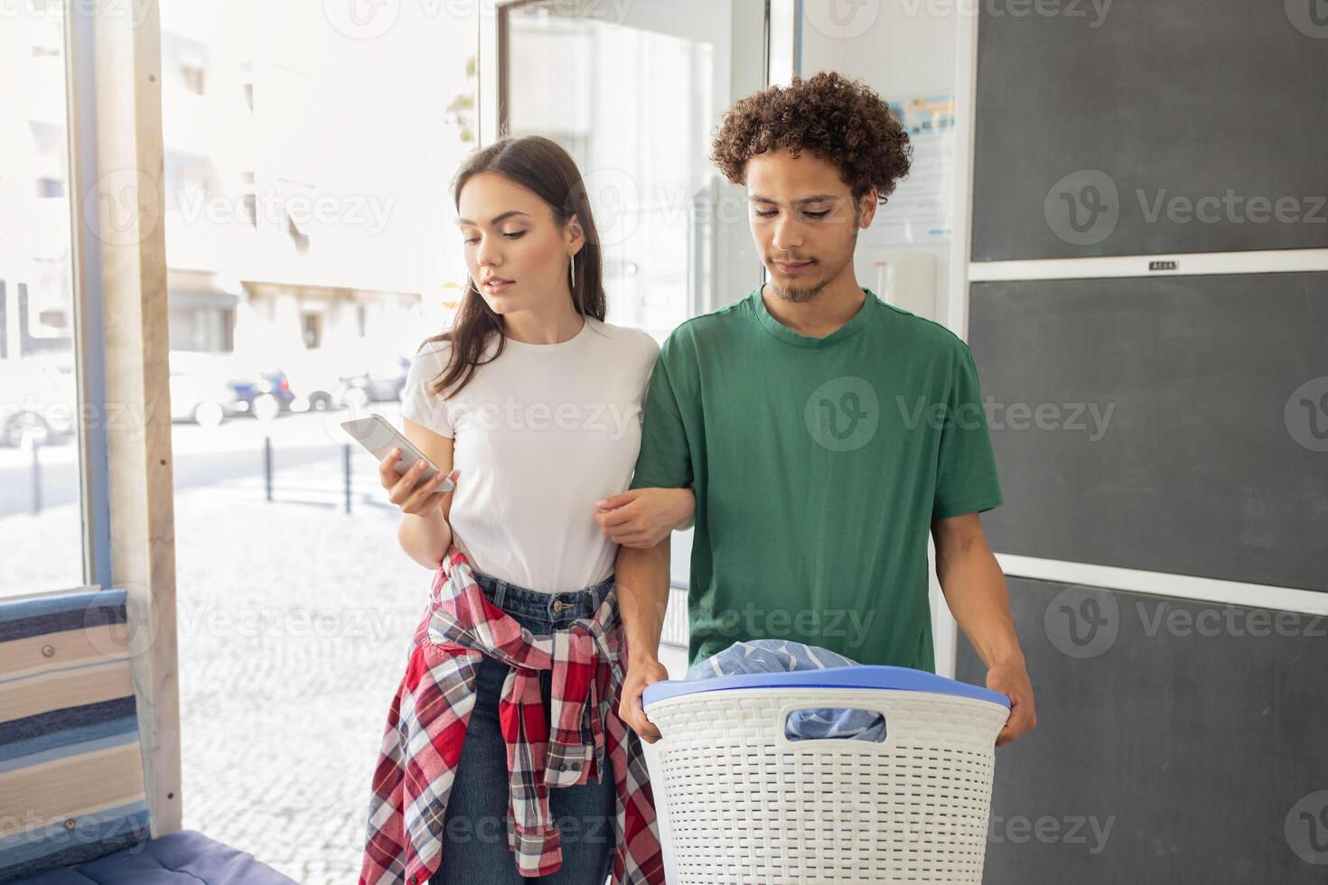 A young couple is working together in a laundromat. One person checks their phone while the other carries a basket of clothes, showcasing teamwork during laundry day. photo