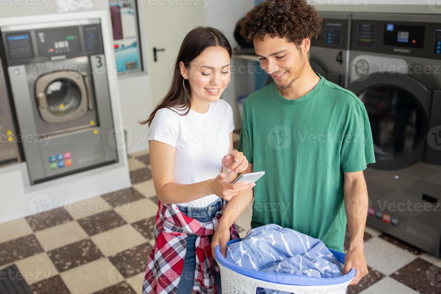 A couple stands together in a laundromat, engaged in conversation while checking their laundry progress on a smartphone, surrounded by washing machines and laundry baskets. photo