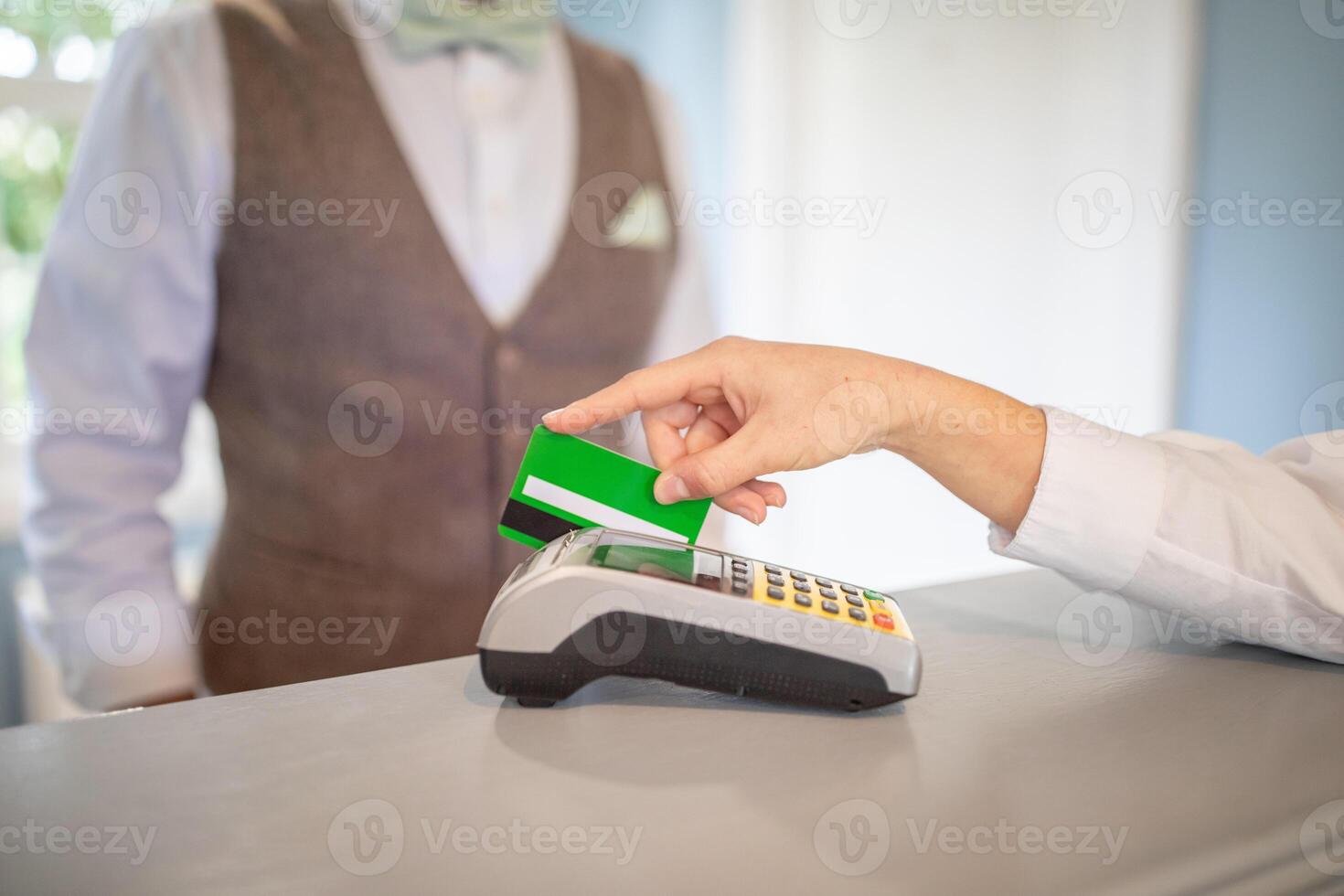 A customer is processing a payment at a retail counter, holding a green card near a card reader, while a staff member stands in a brown vest nearby, ready to assist. photo