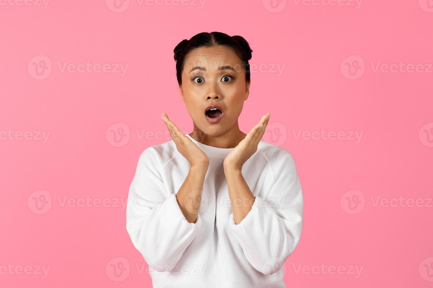 Asian young woman with two playful buns showcases a surprised expression while raising her hands in awe. The striking pink background enhances her joyful demeanor, capturing a moment of delight. photo